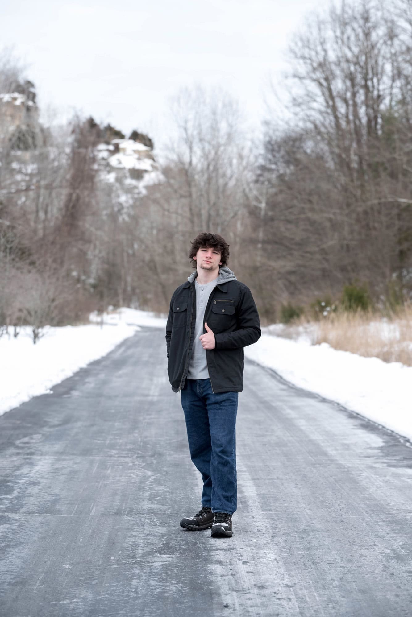 Winter senior photo of a boy wearing blue jeans, a gray henley shirt, and a black jacket on an icy road at Klondike Park.