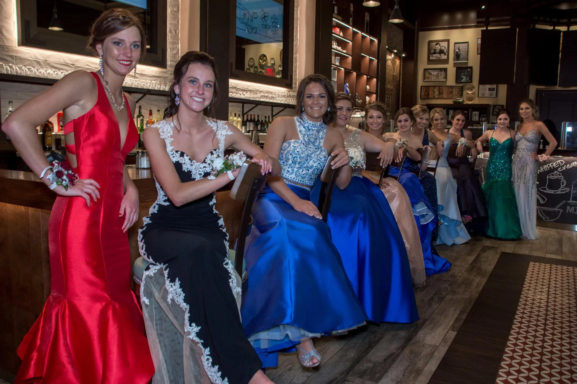 Wide photo of a large group of high school girls in vibrant prom gowns sitting along a wooden bar top inside a St. Louis Missouri restaurant.