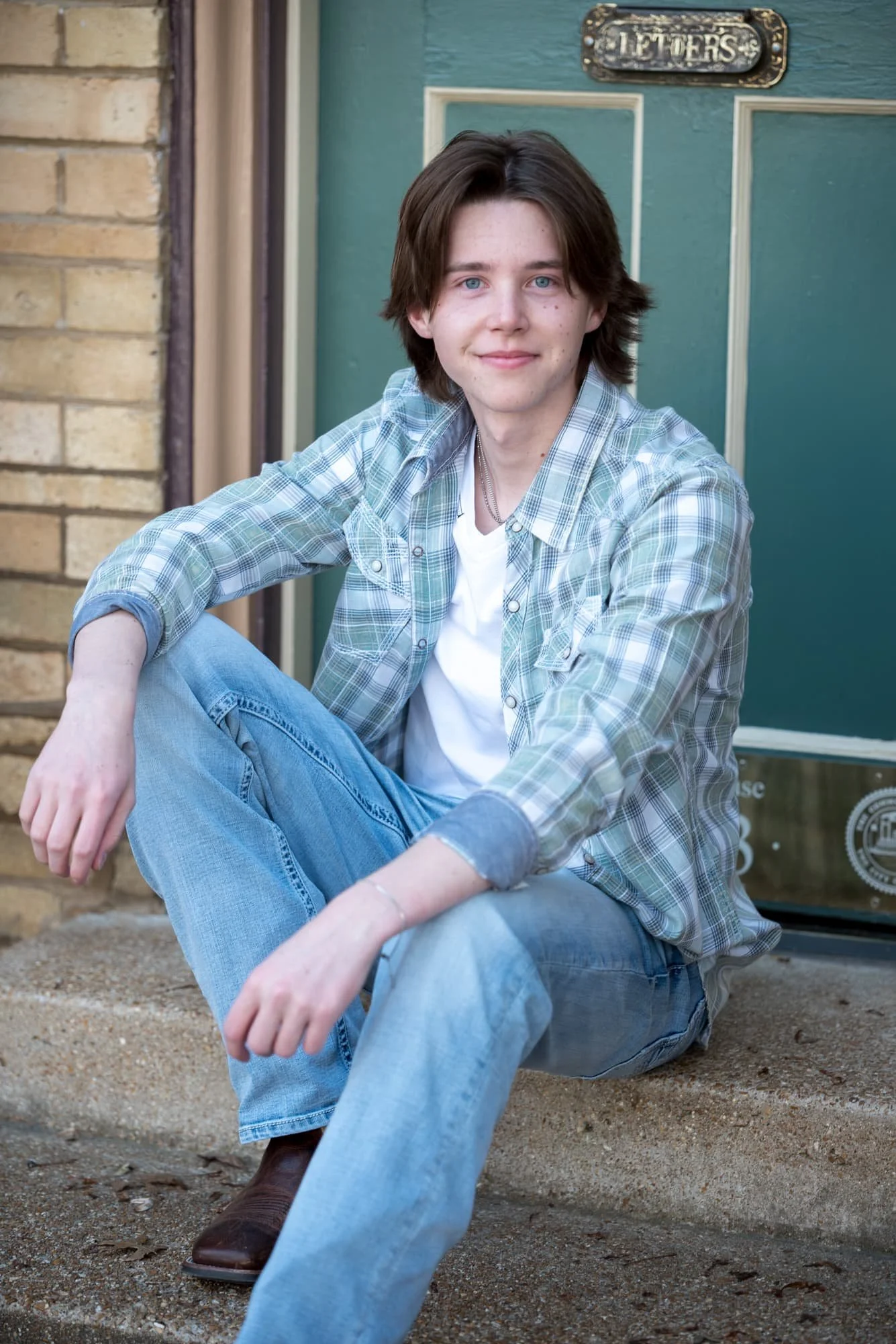 Outdoor senior photo taken in St. Louis, Missouri, featuring a smiling boy in a green and blue plaid shirt, blue jeans, and cowboy boots sitting on the steps of a brick building with a prominent green door.