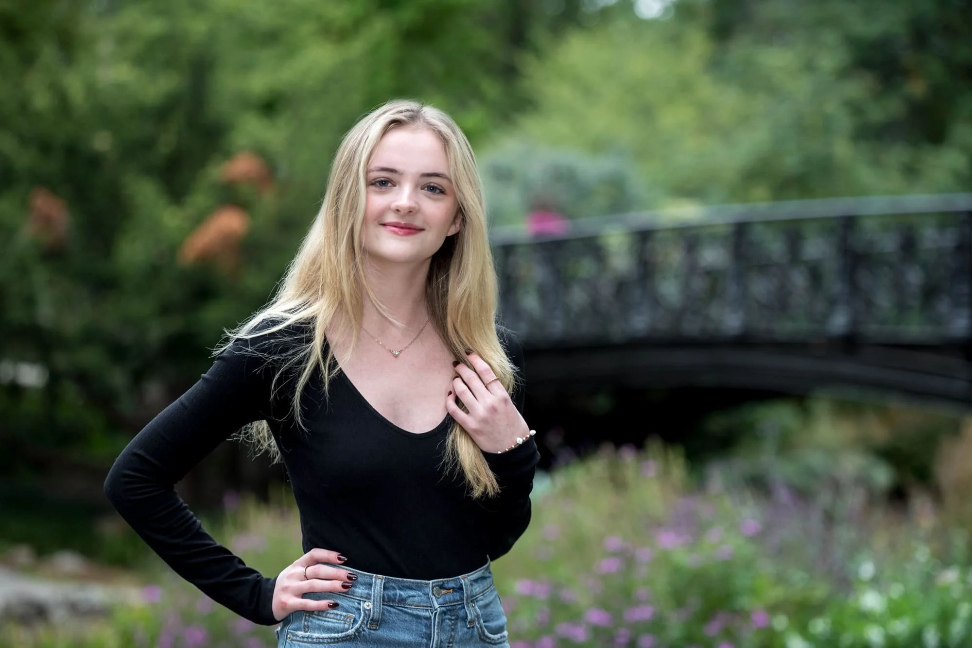 Close-up senior portrait by a St. Louis senior photographer, featuring a girl in a black long-sleeve shirt and blue jeans posing with her hand on her shoulder, framed by a historic black bridge and vibrant purple flowers.