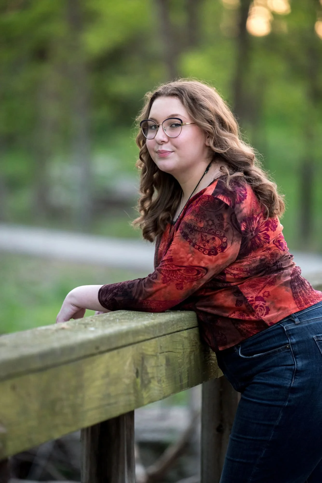 A spring sunset picture captures a high school senior girl in a red shirt and blue jeans standing on a bridge in the woods for a photo by a West County photographer.