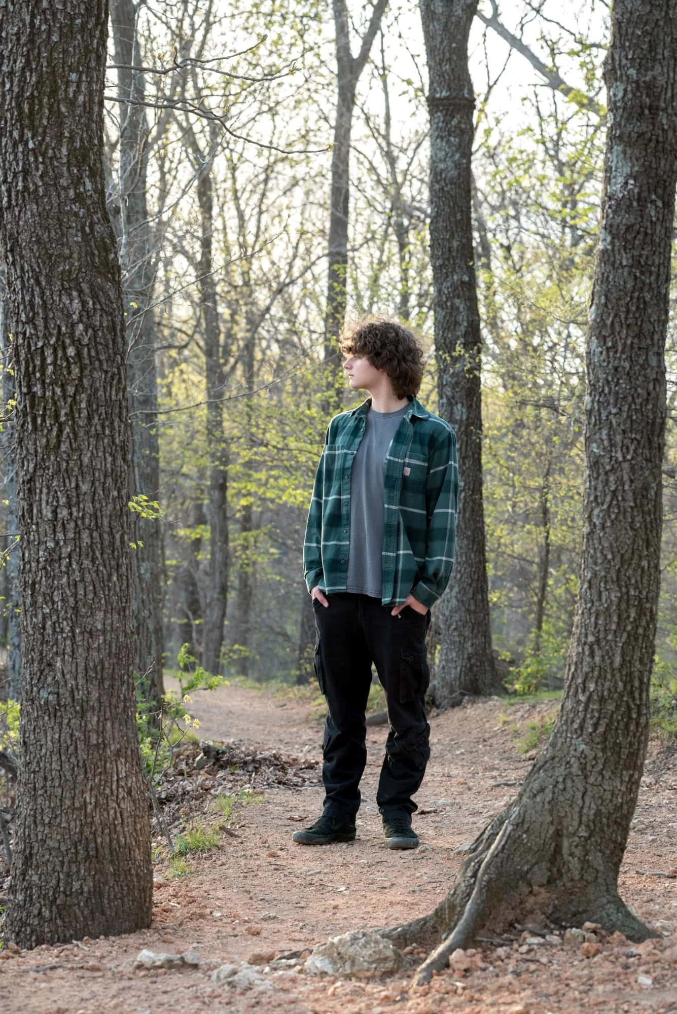 Senior picture of a guy looking off into the distance, standing on a wooded pathway in early spring wearing a green plaid long-sleeve and black pants at Castlewood State Park.