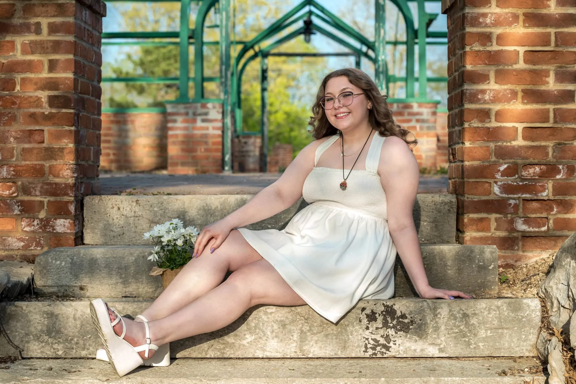 High school senior girl wearing an elegant white dress and white wedges, smiling while sitting on the stone steps in front of the historic green greenhouse structure at Queeny Park in St. Louis.