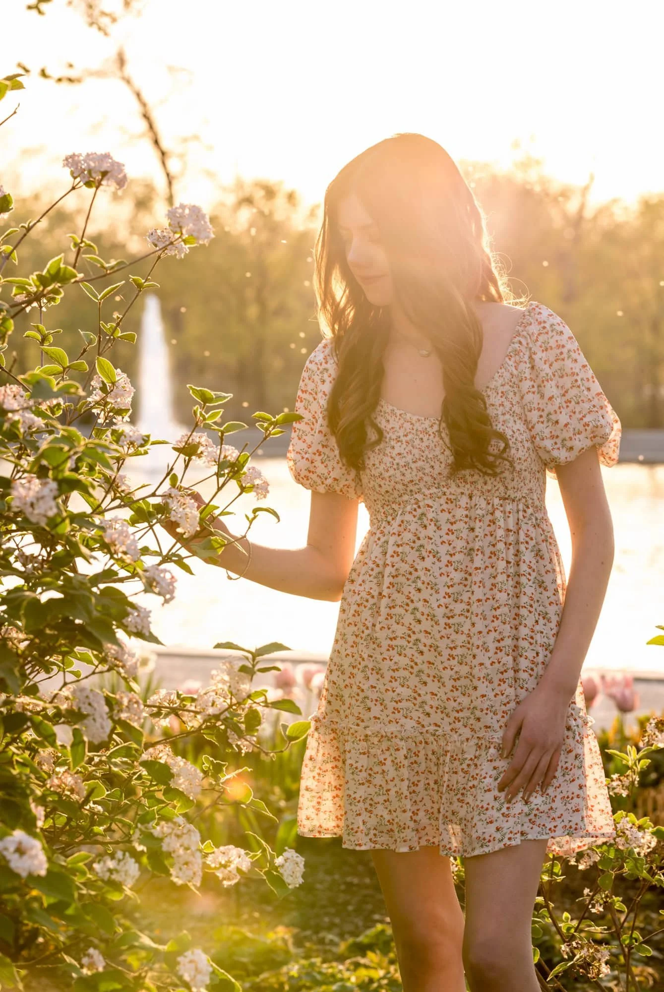 Whimsical senior picture of a girl wearing a white dress with red flowers, holding a spring flower bush in her hand as dramatic sunset backlighting streams into the frame.