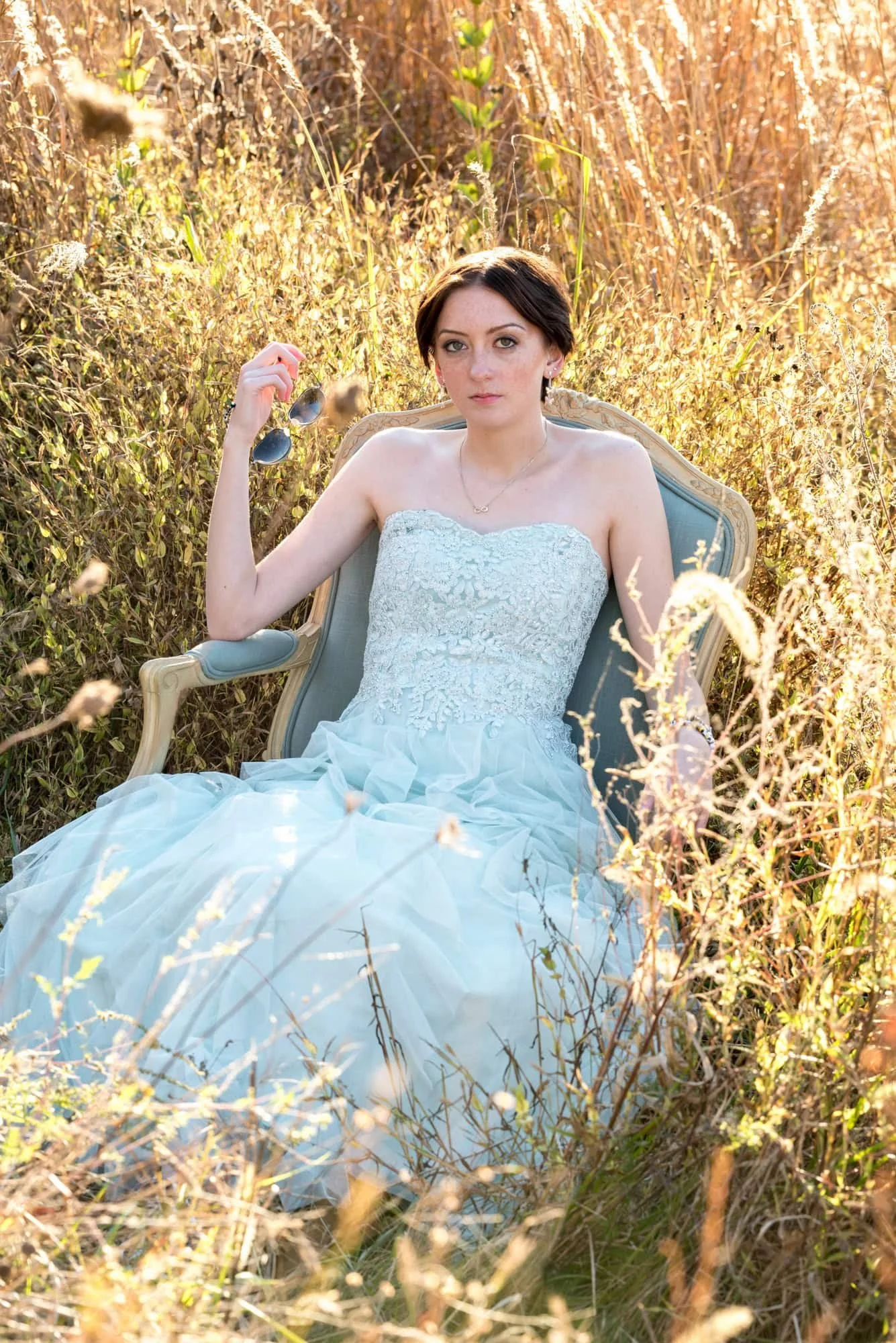 Stunning outdoor senior photo of a girl with a serious expression wearing a mint green prom dress, sitting in a blue chair in the middle of a sunlit field of tall grass at Queeny Park.