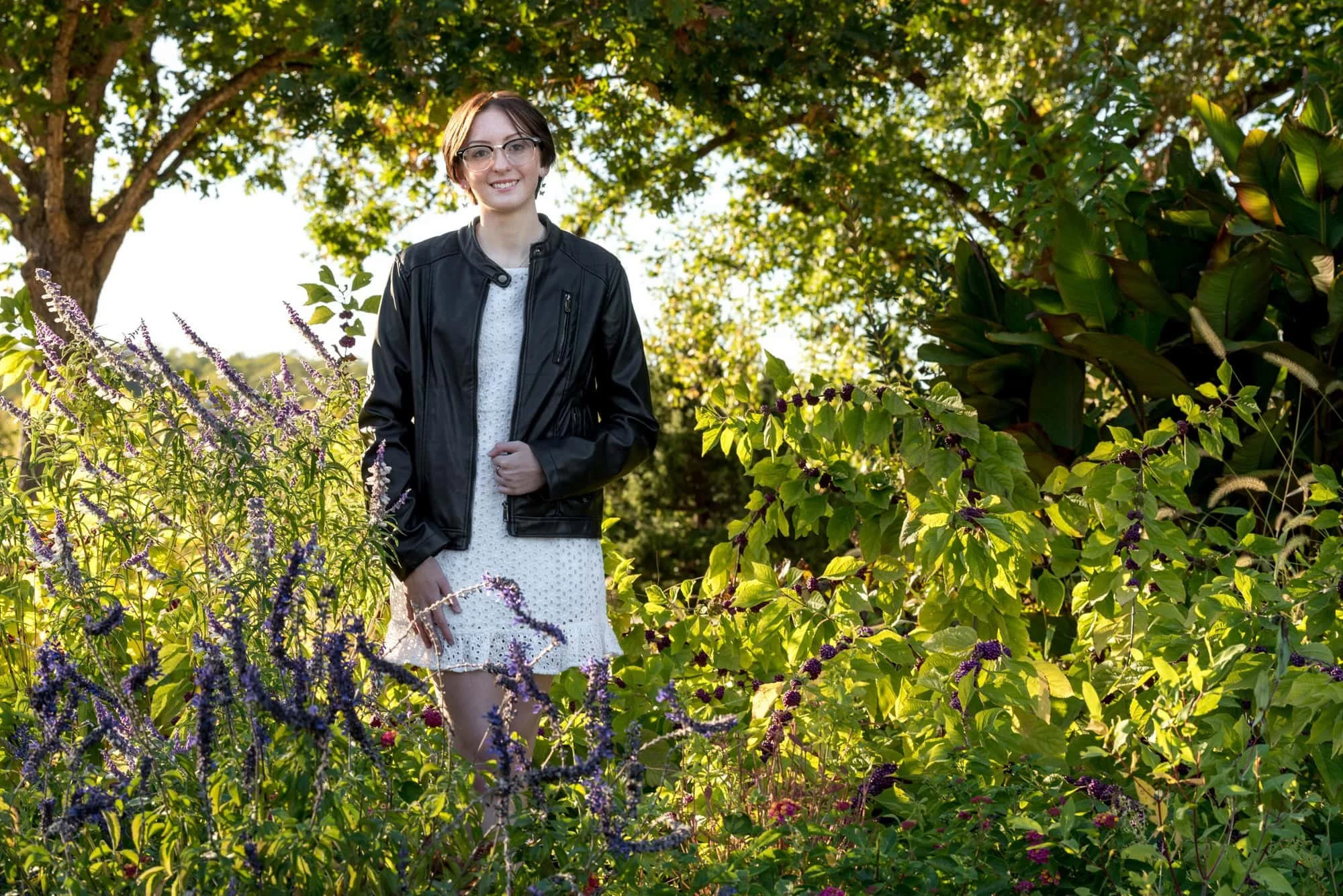 Vibrant outdoor senior picture showing a girl posing in a black leather jacket and a white dress, standing in the middle of lush greenery and purple flowers at Queeny Park.