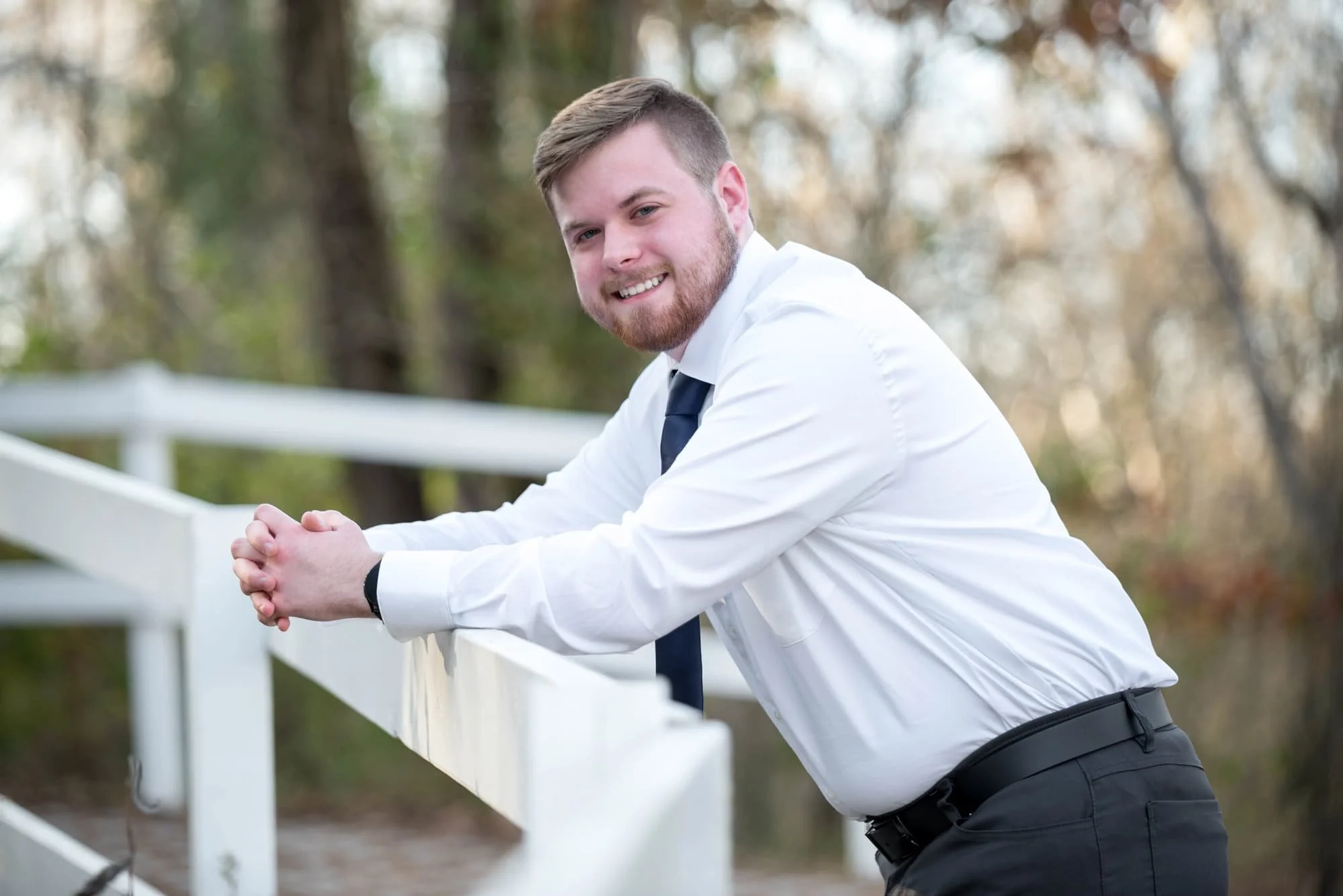 Leaning against a wood fence during a fall session in Hillsboro, Missouri, a senior guy wearing a white button-up shirt and navy blue tie poses for a picture.