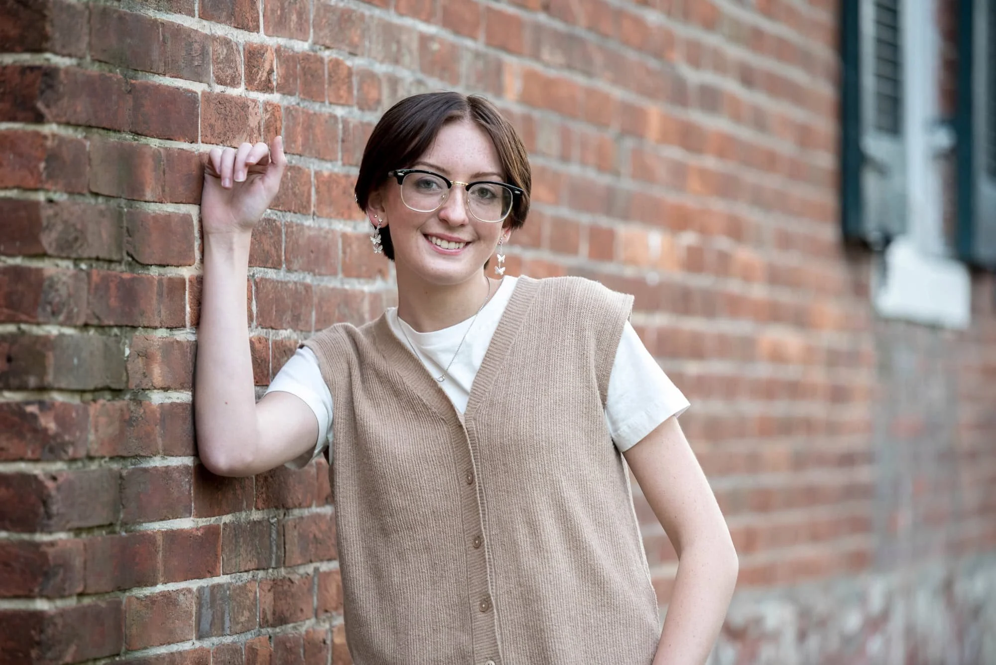 Classic architectural senior picture featuring a girl wearing a tan vest and white t-shirt, resting her arm against the textured brick wall of the historic Victorian house at Queeny Park.