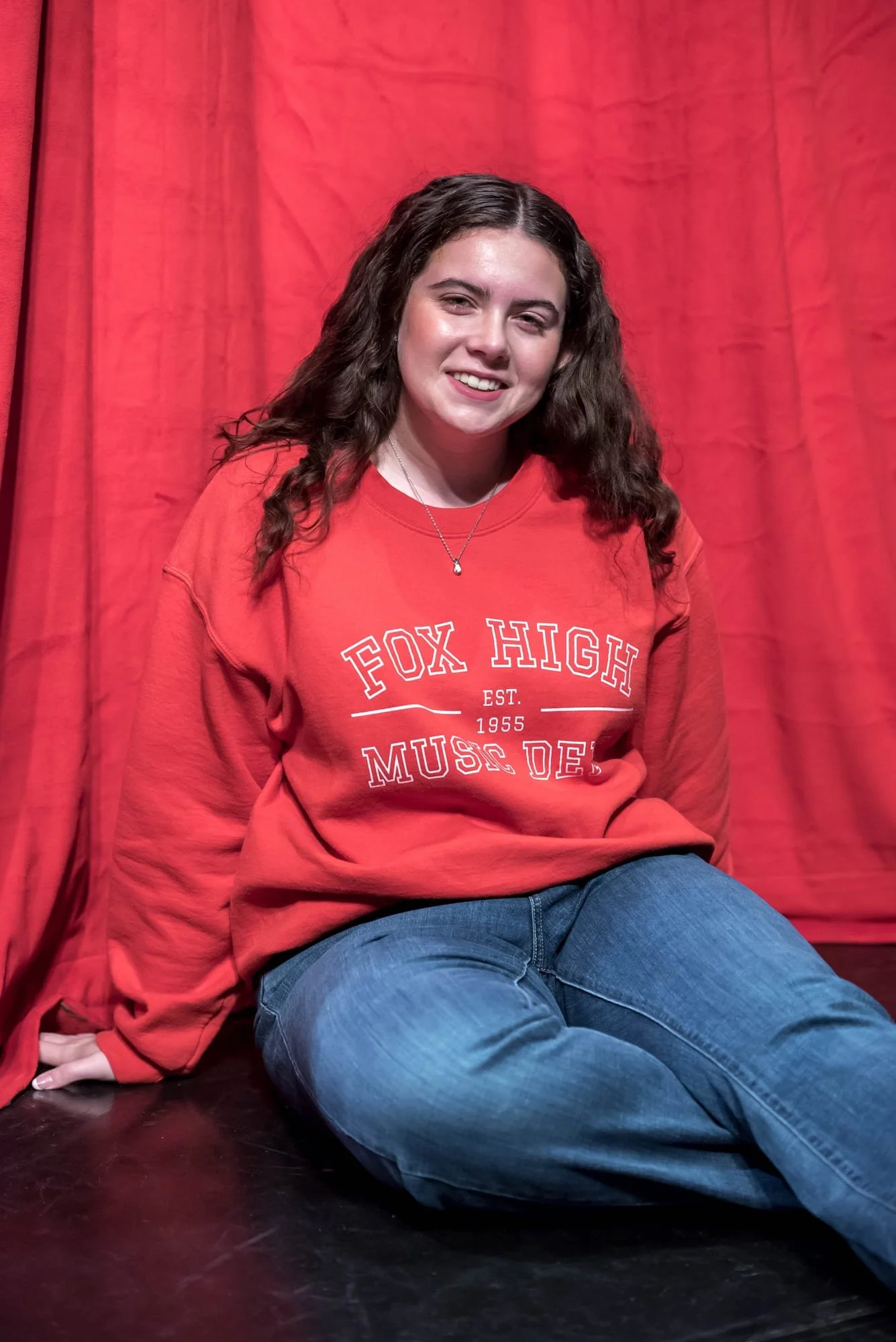 Sitting on the auditorium stage in Arnold, Missouri, a senior girl in a red music department sweatshirt and blue jeans poses for a picture.