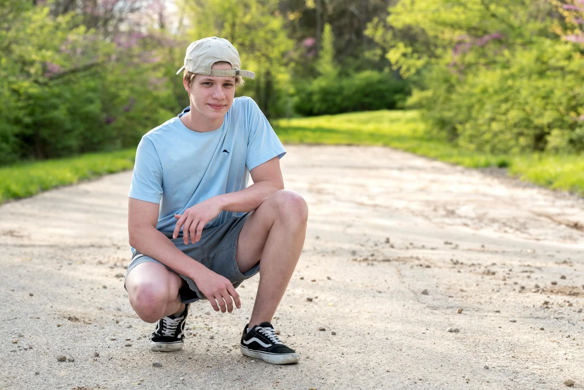Casual senior picture of a guy wearing a backwards hat, light blue t-shirt, and grey shorts with black shoes, squatting down on a quiet dead-end road bathed in bright spring sunlight in Columbia IL.