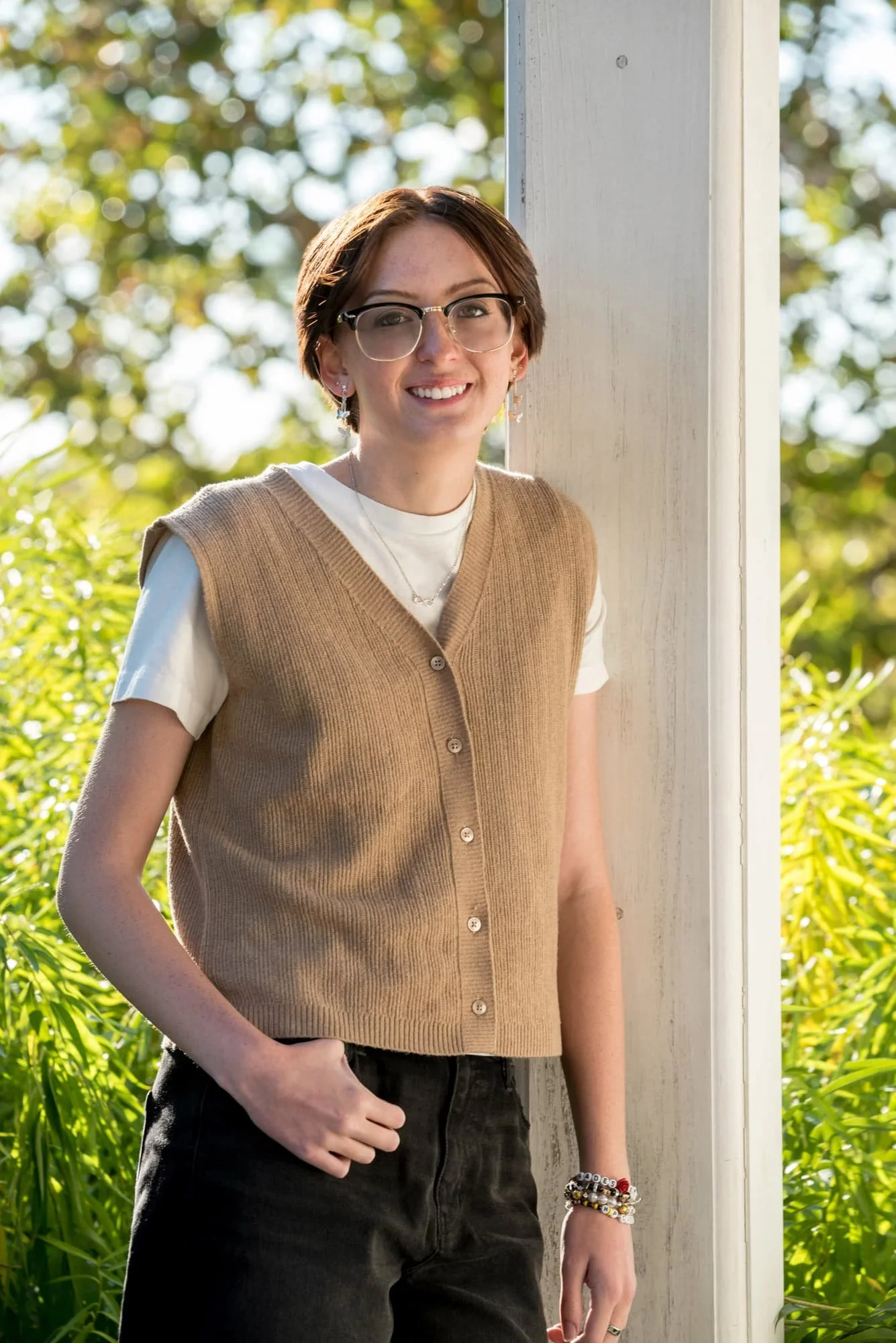 Casual park senior picture of a girl wearing a tan vest, white t-shirt, and black jeans, leaning on a white pillar in front of sunlit green bushes at Queeny Park.