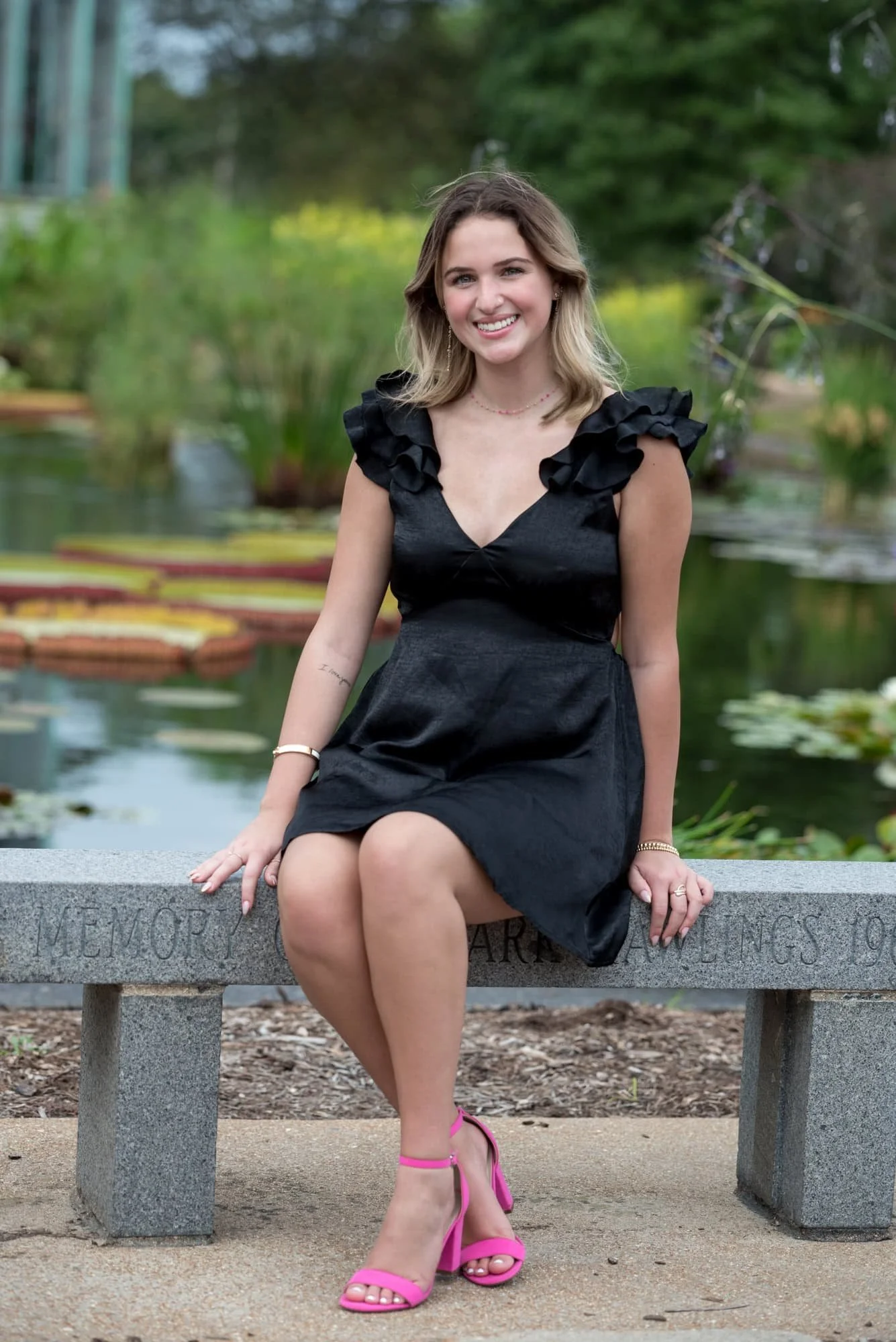 Picturesque outdoor image featuring a senior in a black dress and pink heels sitting on a stone bench beside a lily pad pond at the Jewel Box.