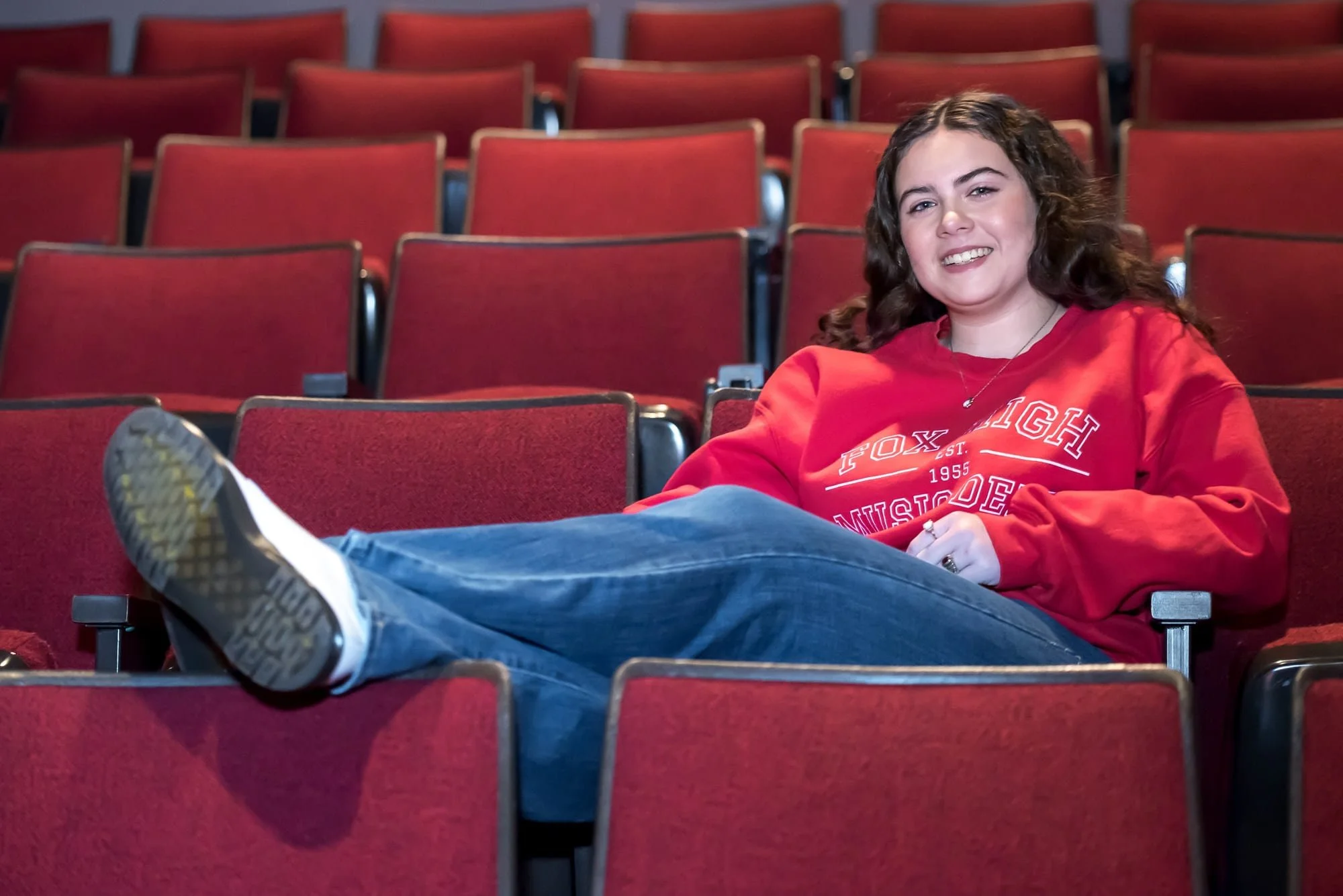 High school senior girl in a red Fox High School music department sweatshirt, sitting relaxed in the red theatre seats of Rickman Auditorium with her feet kicked up on the rows in front of her.