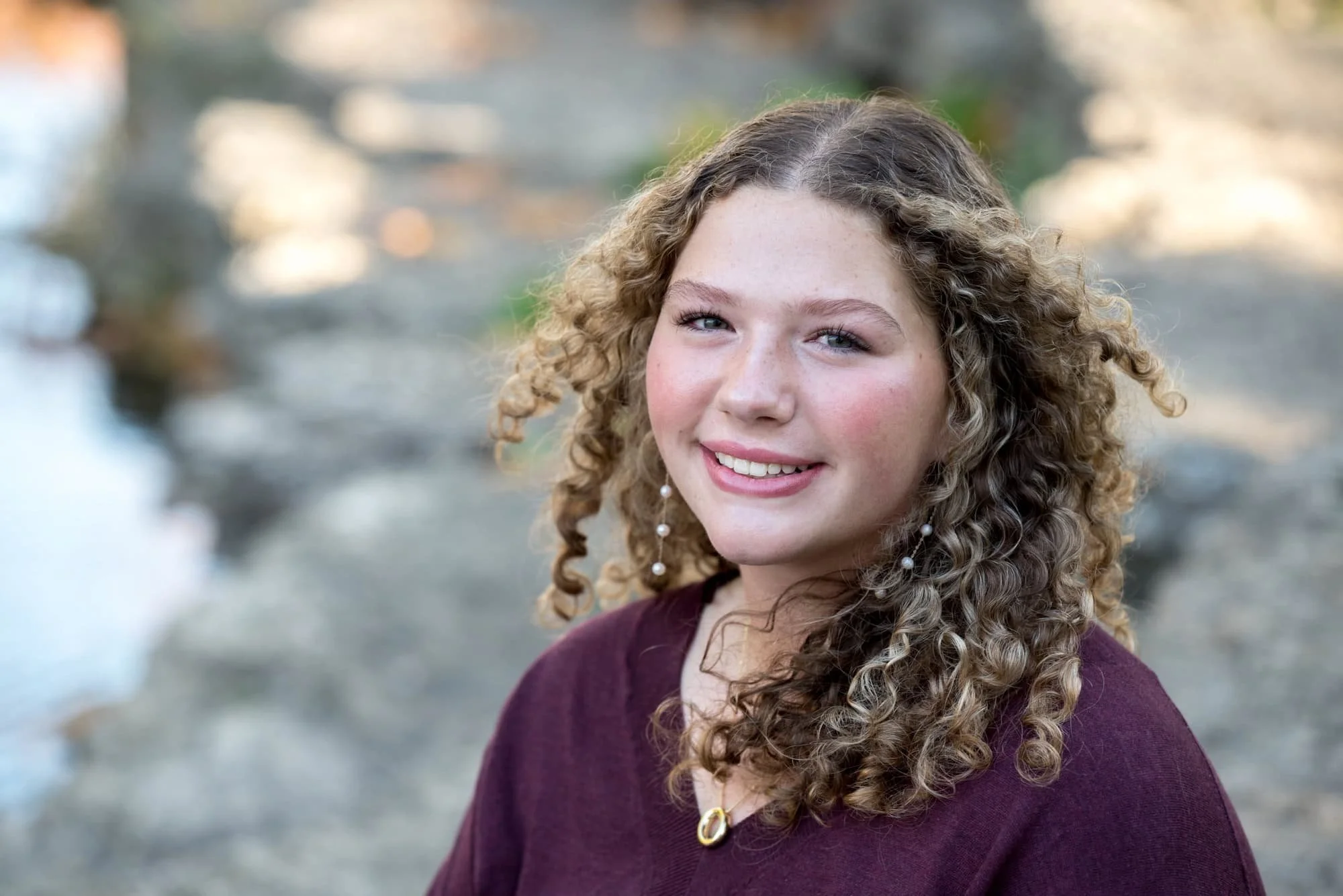 Close-up high school senior picture showcasing a smiling girl wearing a purple shirt with her curly hair blowing in the wind near The Muny.