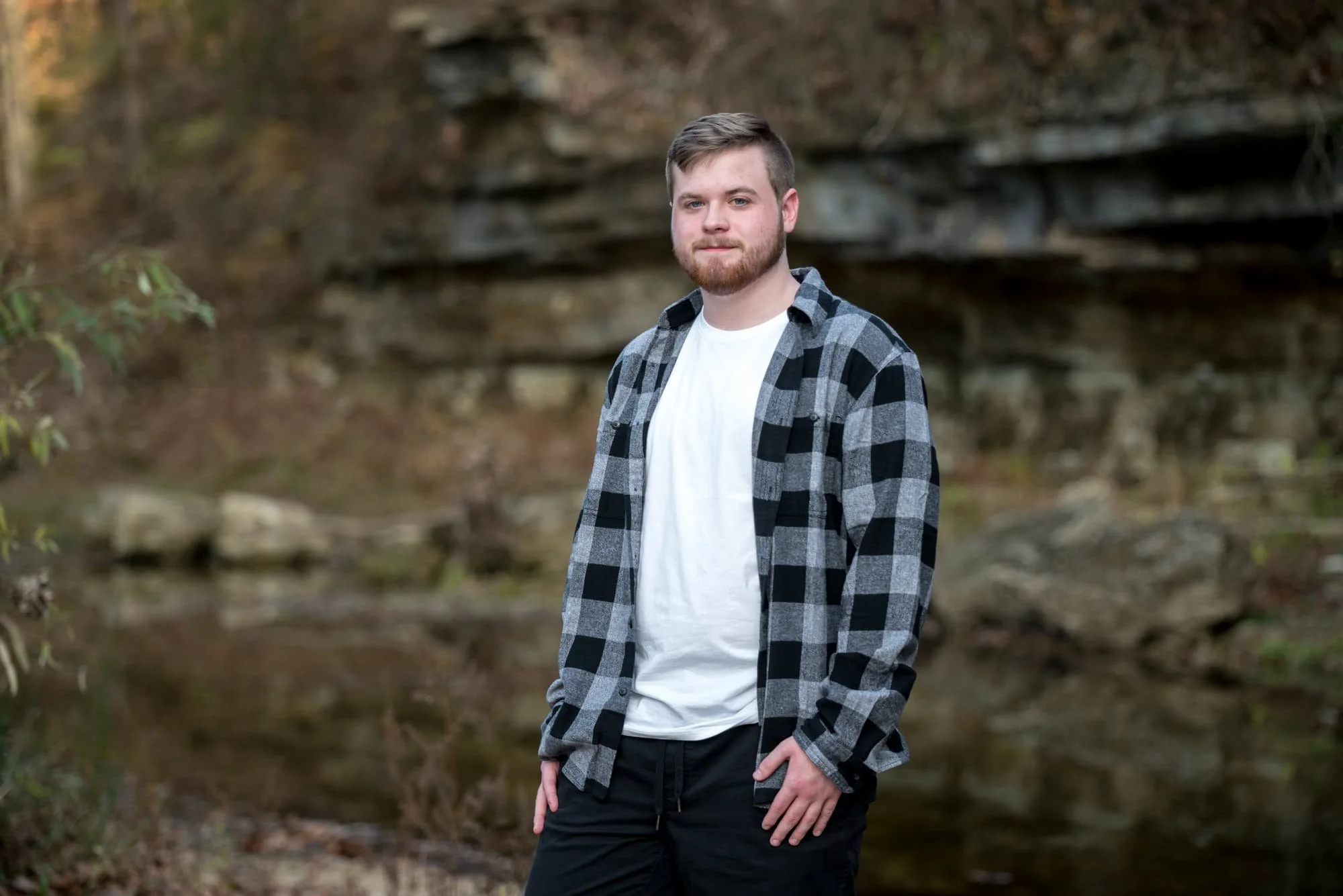 Along the rocky edge of Sandy Creek, a senior guy wearing a plaid shirt and black pants poses for an outdoor picture.