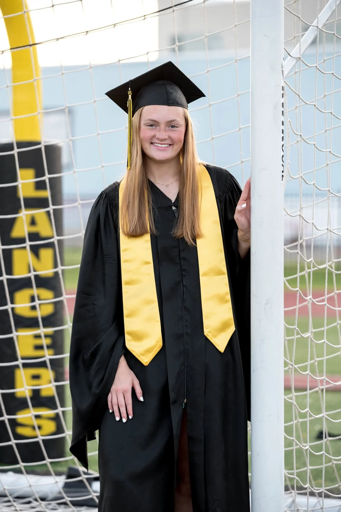 Standing outdoors in Affton, a senior girl in a black graduation cap and gown with a gold stole poses for a picture in the goalie net.
