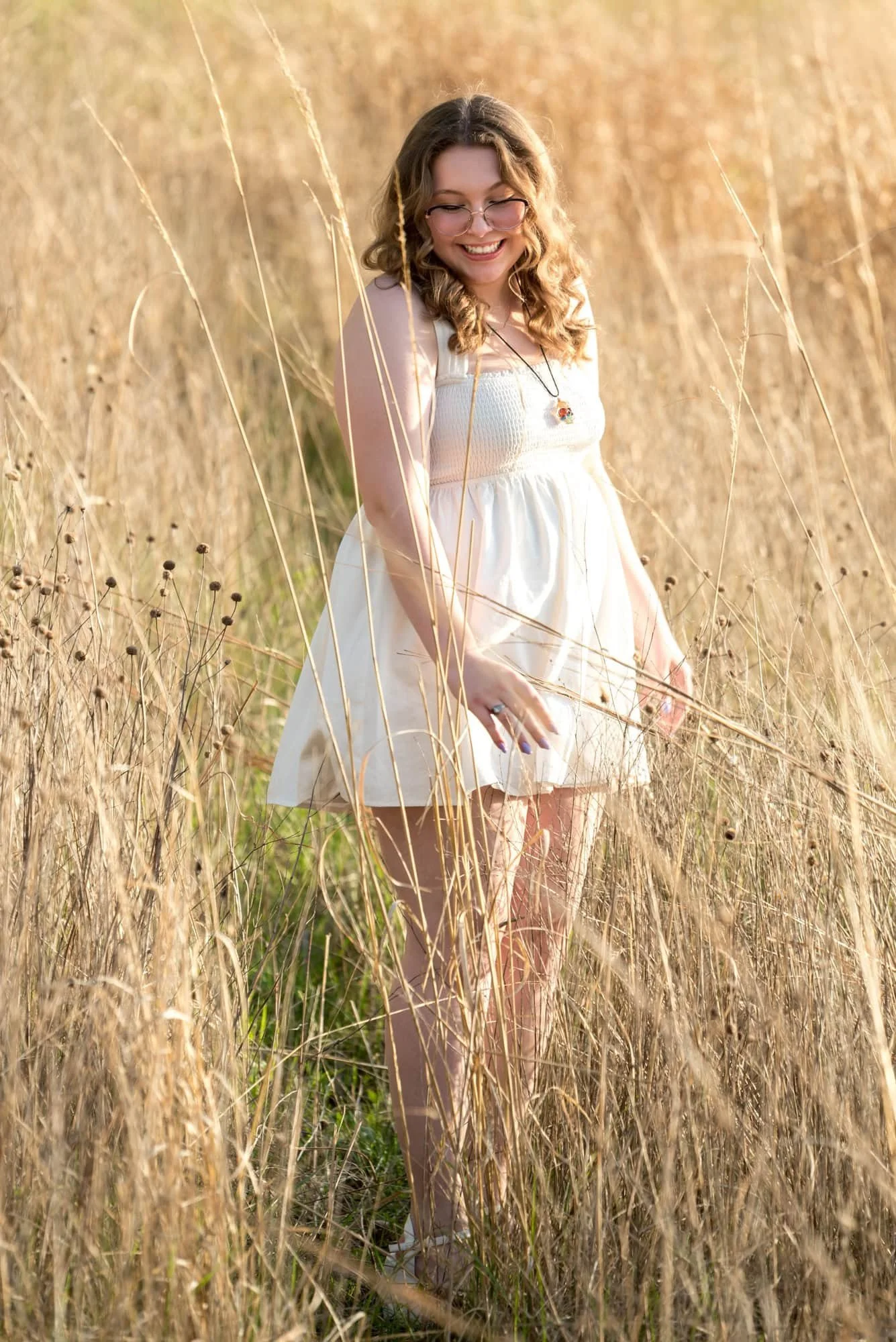 Senior girl in a flowing white dress, smiling as she twirls and looks down in a sun-bathed tall grass field during a golden hour portrait session at Queeny Park.