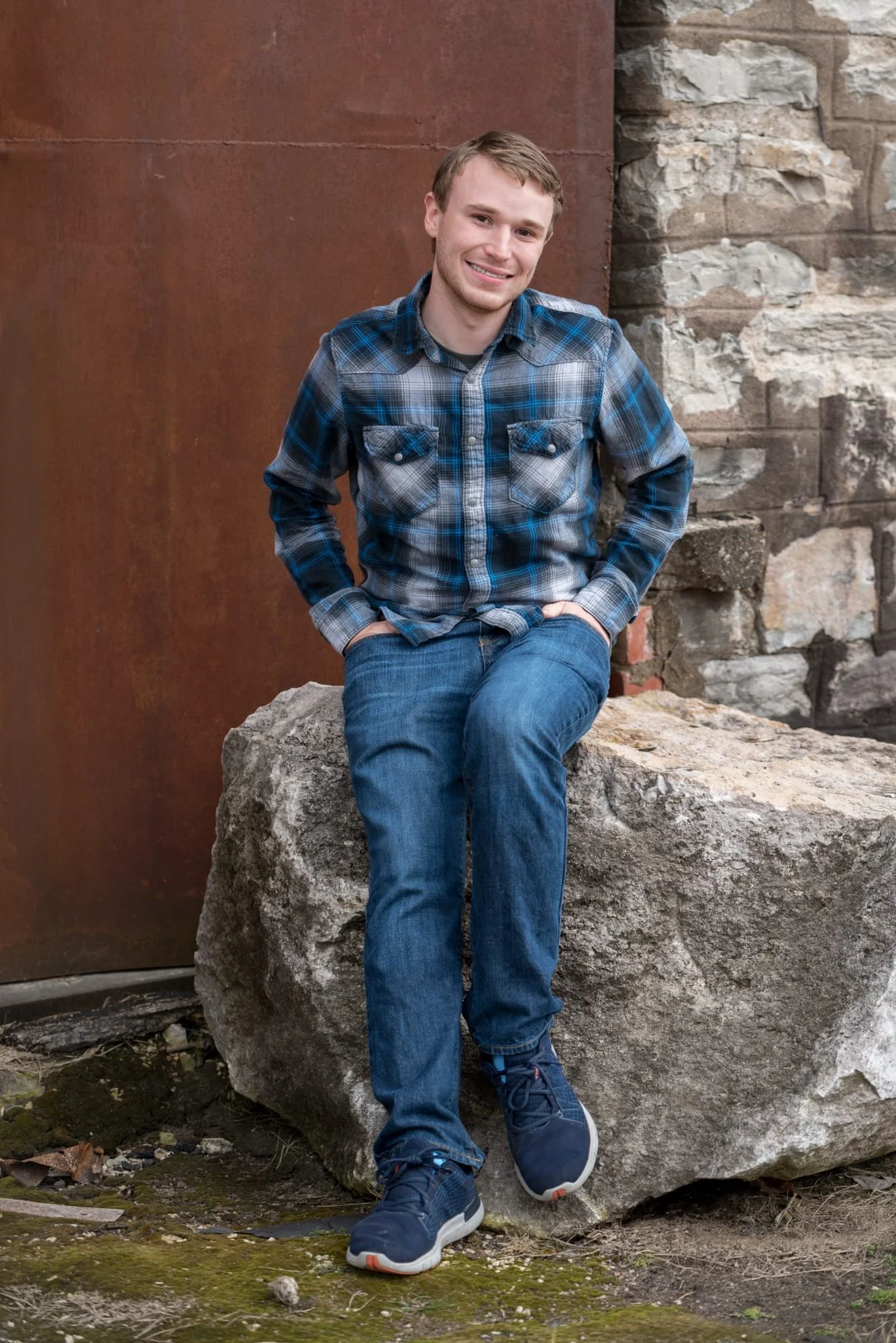 High school senior guy wearing a blue plaid shirt and jeans, posing on a rock in front of a weathered rusty door and historic stone building at Rock City in Valmeyer IL.