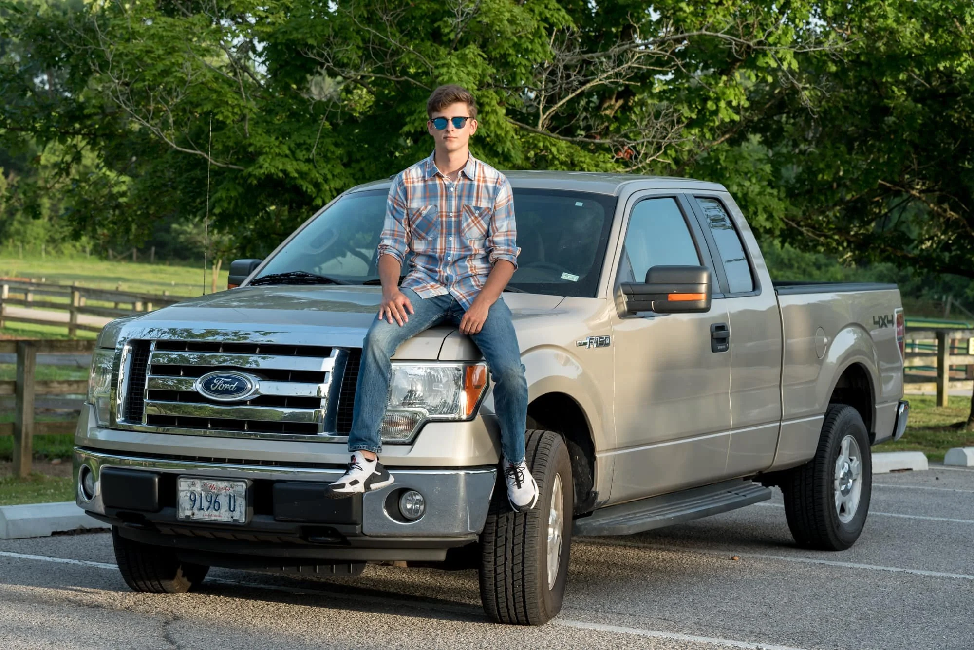 Casual senior guy portrait of a student wearing sunglasses, an orange and blue plaid shirt, and blue jeans, sitting coolly on the hood of his Ford F-150 truck at Suson Park.