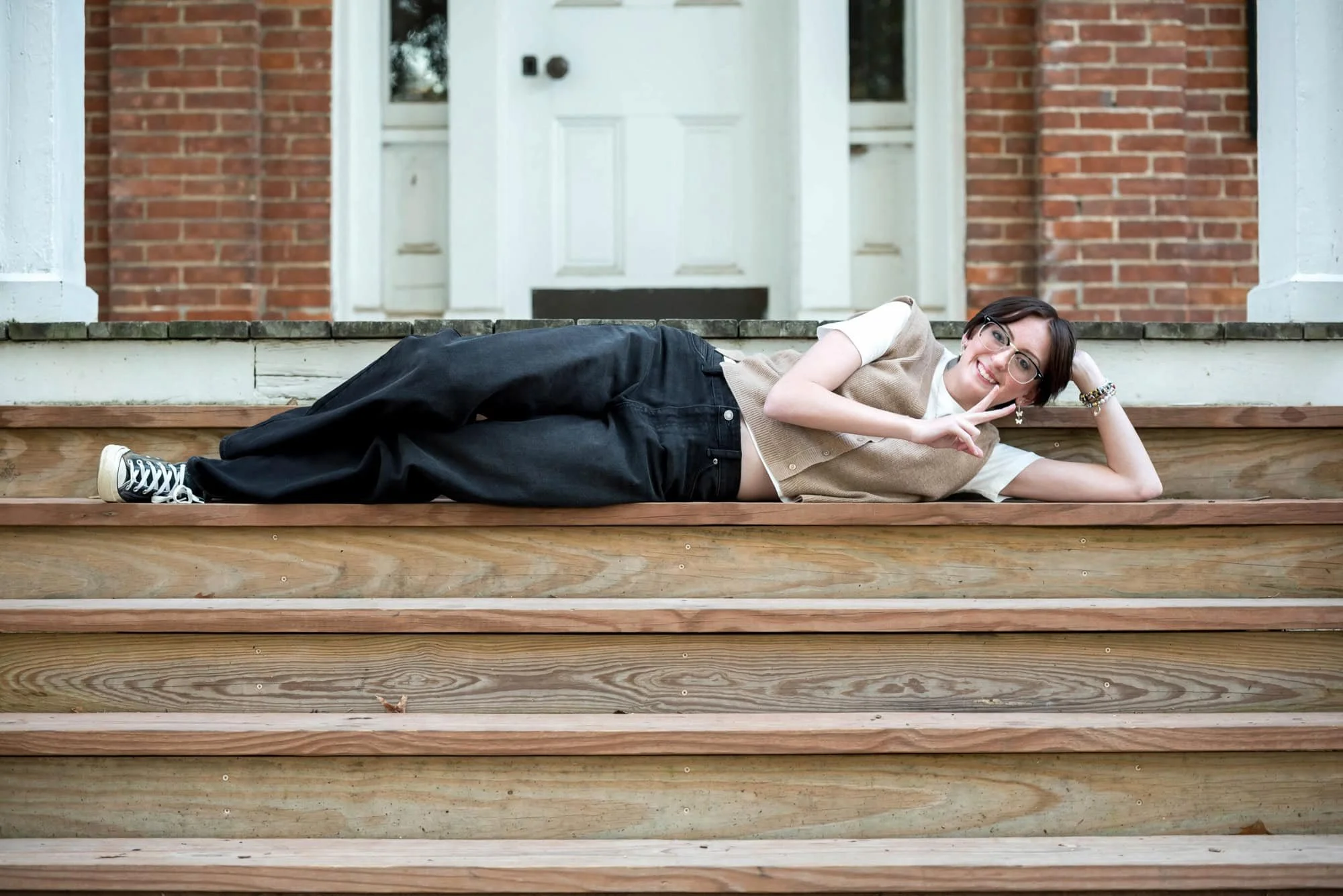 Casual park senior photo of a girl flashing a peace sign while laying on her side on the wooden steps of the Victorian house at Queeny Park, wearing a tan vest, white t-shirt, and black pants.