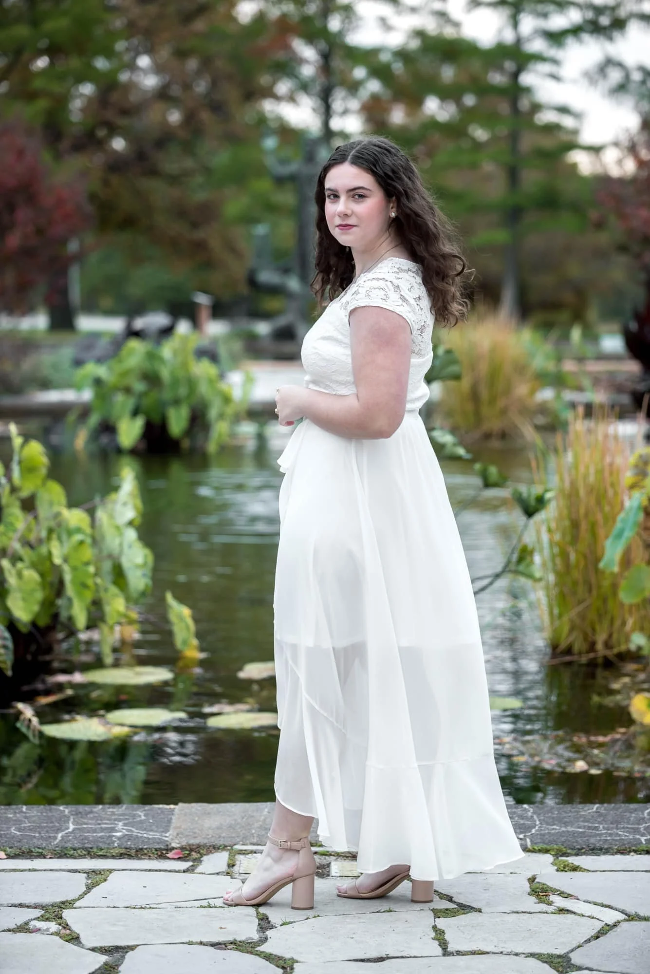 A fall picture features a high school senior girl wearing a white dress and beige heels standing by a pond on a stone pathway at the Jewel Box.