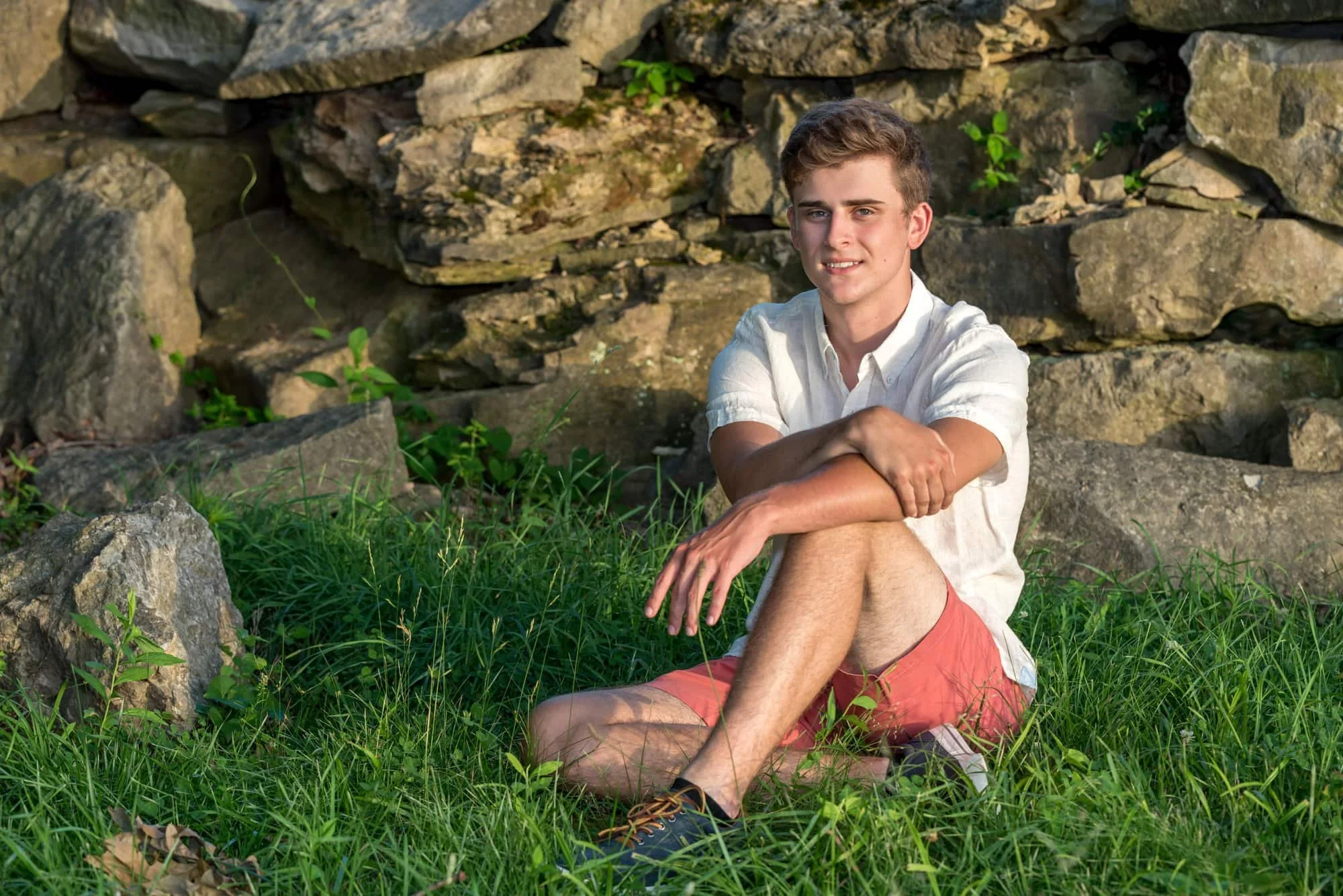 Classic and casual senior picture of a guy wearing a crisp white button-up shirt, orange shorts, and grey shoes, sitting relaxed in the grass with his arm resting on his knee in front of a rustic rock wall in Oakville.