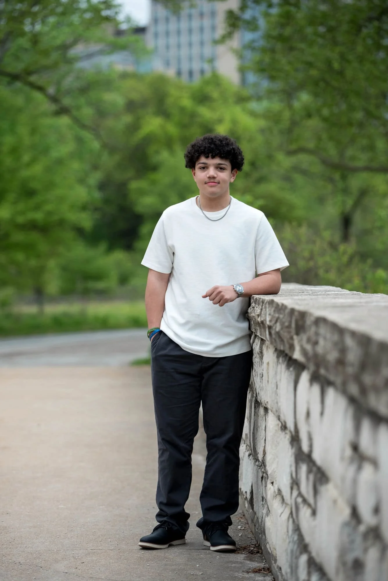 Senior picture of a guy standing on a stone bridge with historic buildings and lush trees in the background at Forest Park.