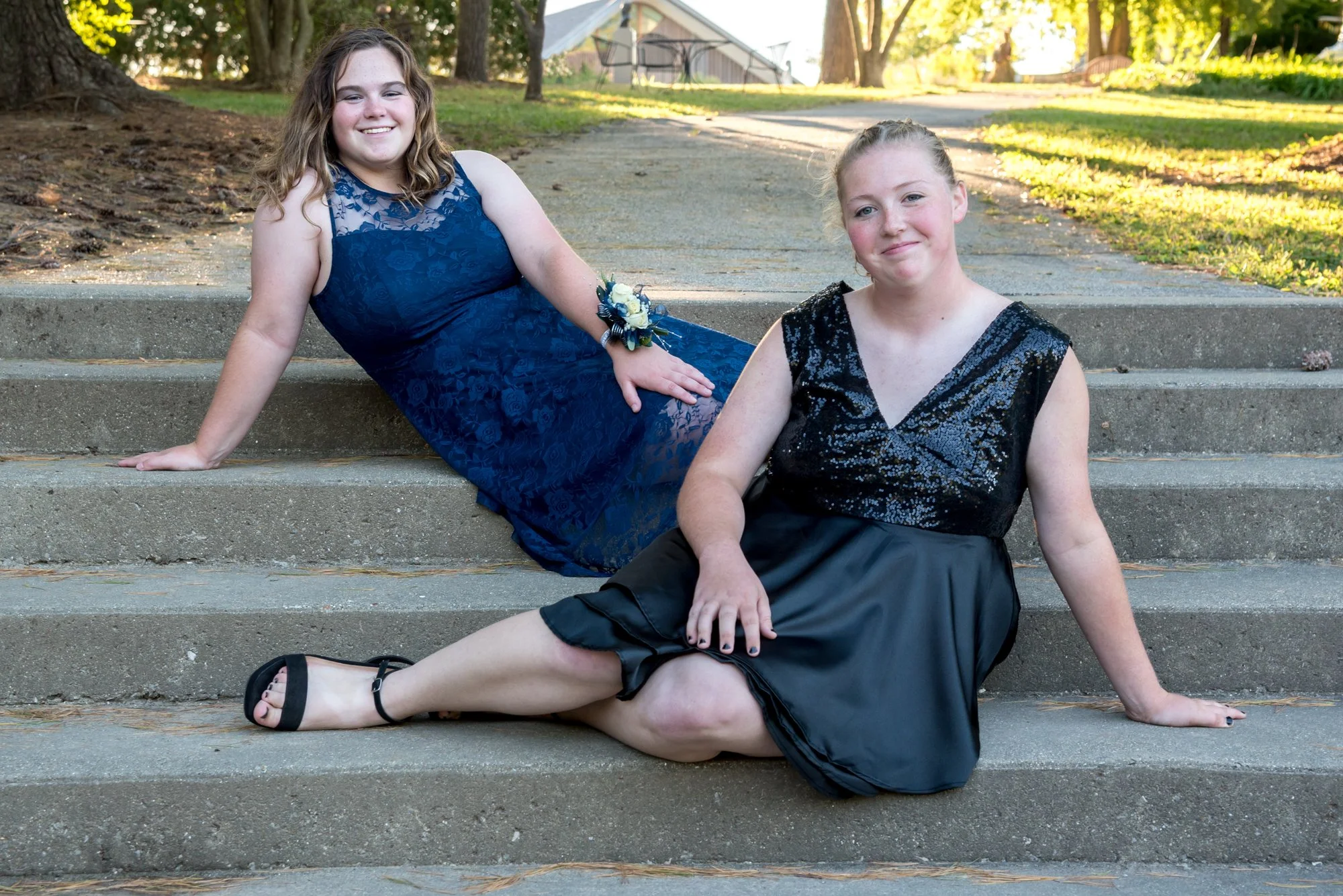 Lovely sun-drenched outdoor photo of two best friends in their formal homecoming dresses, playfully lying across wide park steps with one in front of the other at Laumeier Sculpture Park in Sunset Hills.