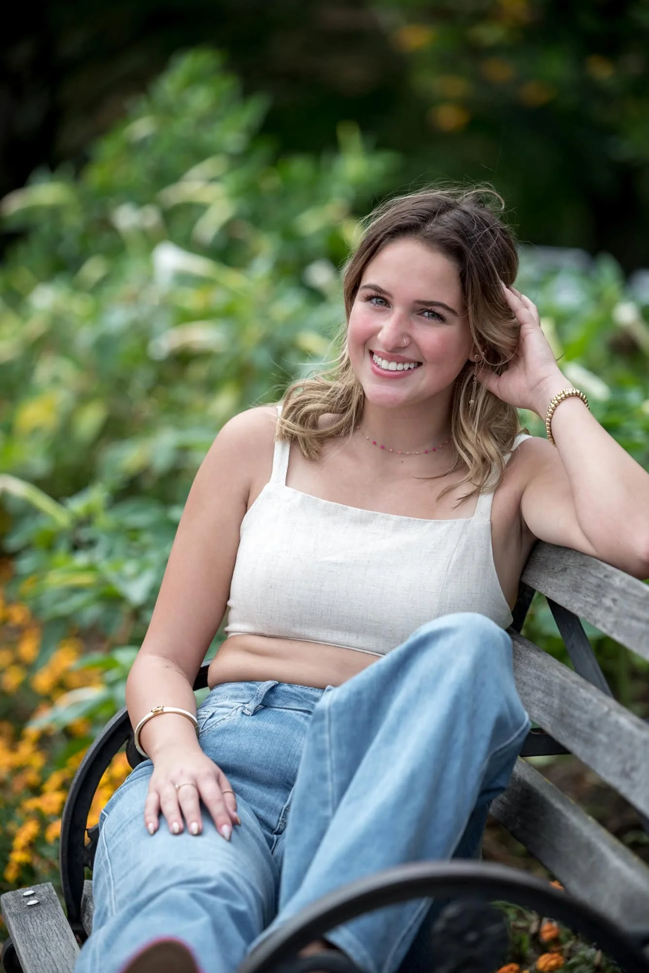 Relaxed summer senior picture at the Jewel Box showing a girl in a gray crop top and blue jeans sitting on a wooden bench surrounded by green foliage.