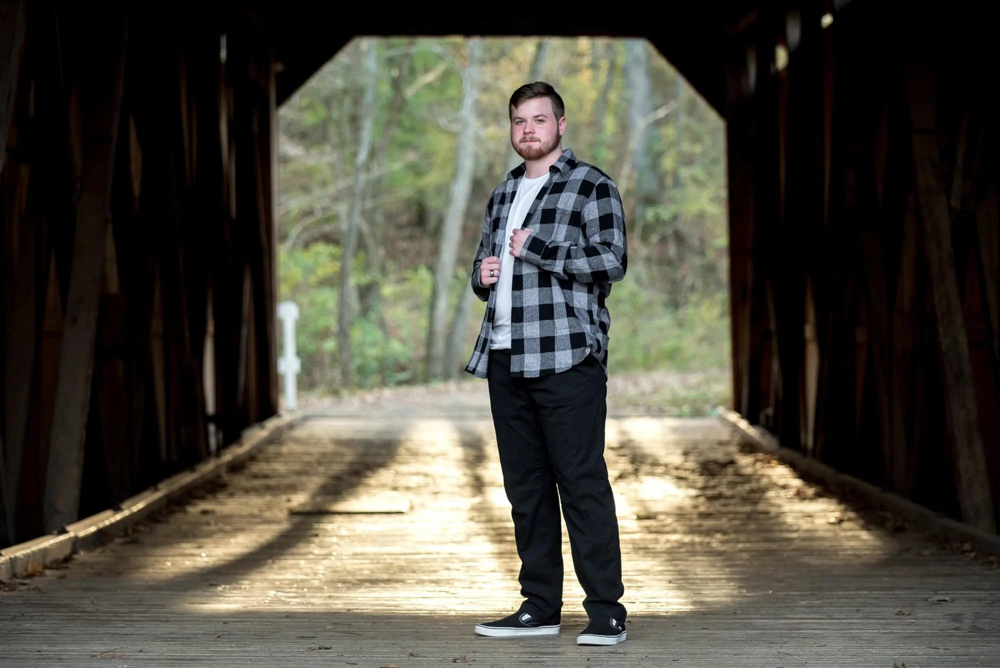 Moody and rustic senior portrait of a guy wearing an open black plaid shirt over a white t-shirt and black pants, standing inside a historic wooden covered bridge over a sandy creek with warm sunlight filtering through the background.