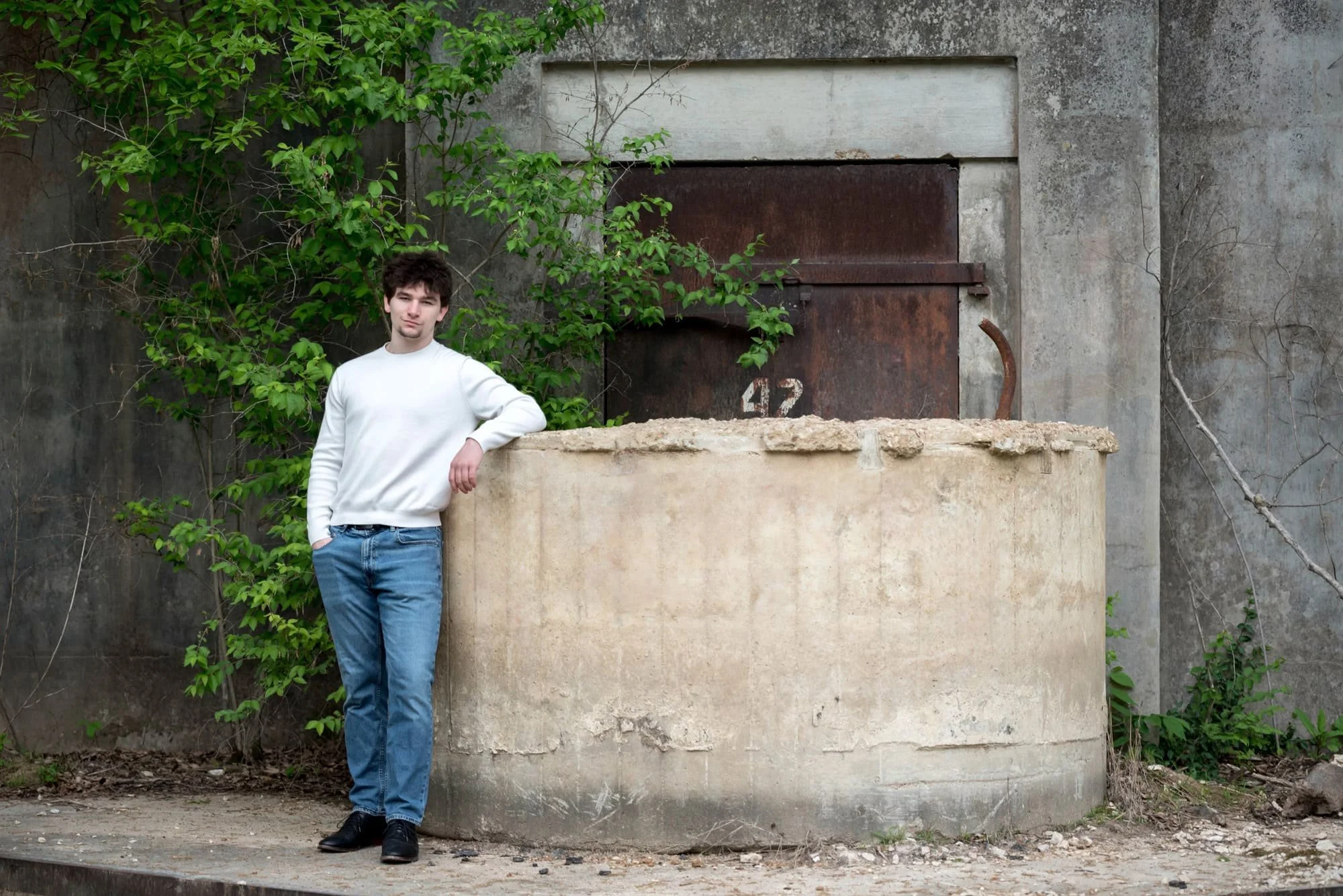 A picture of a senior guy in a white crewneck sweater leaning on a concrete structure in front of bunker number 42 at the August A. Busch Memorial Conservation Area.