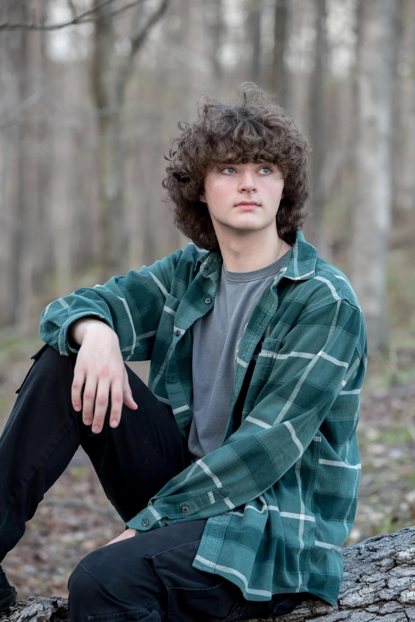High school senior picture of a guy sitting on a log in the middle of the woods, looking off into the distance with his arm resting on his leg for a peaceful, rustic nature portrait by West County senior photographer.