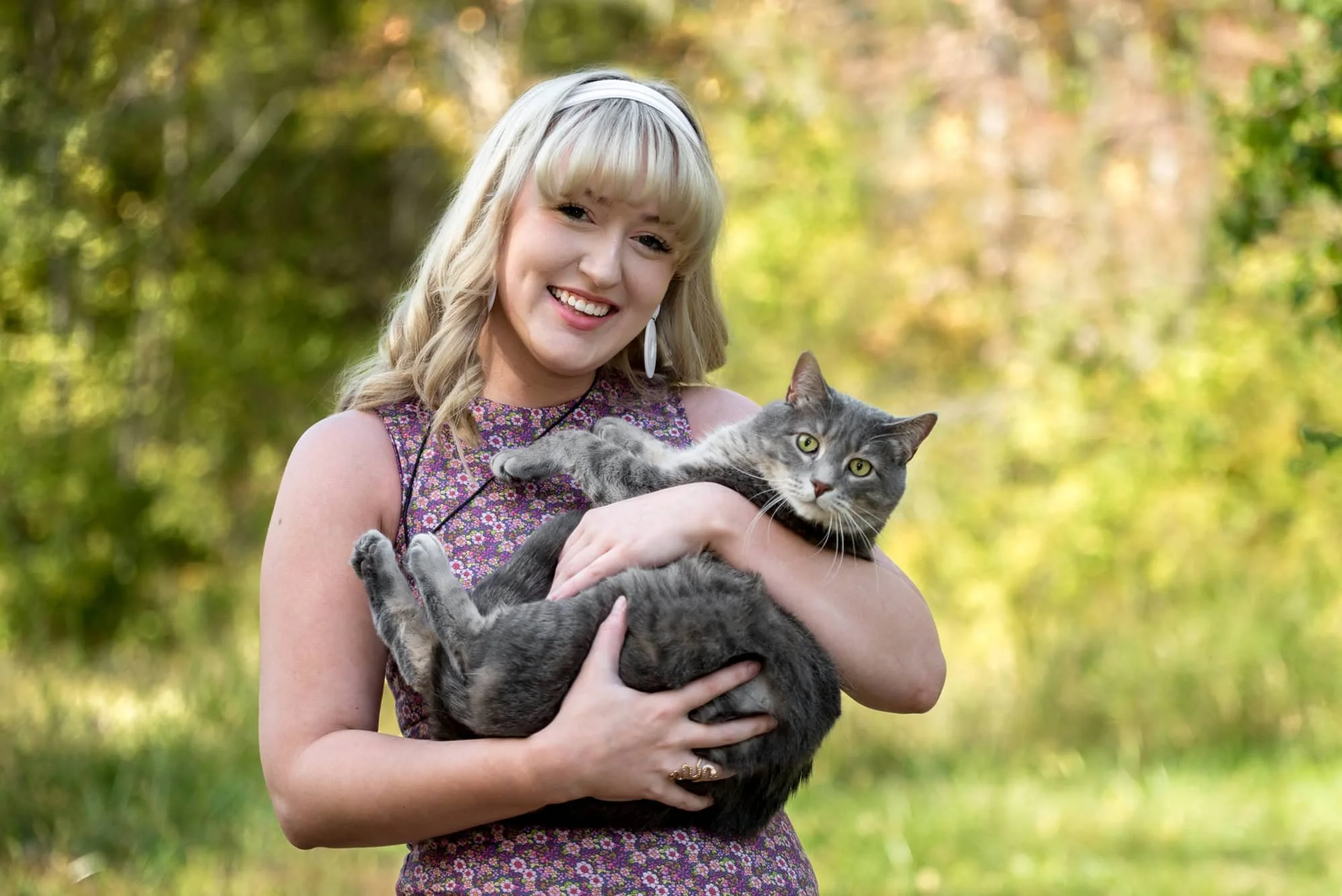 Sweet senior photo by a Columbia IL senior photographer, showing a girl in a purple floral dress and a white headband cradling her grey cat in front of the woods during a glowing golden hour sunset.