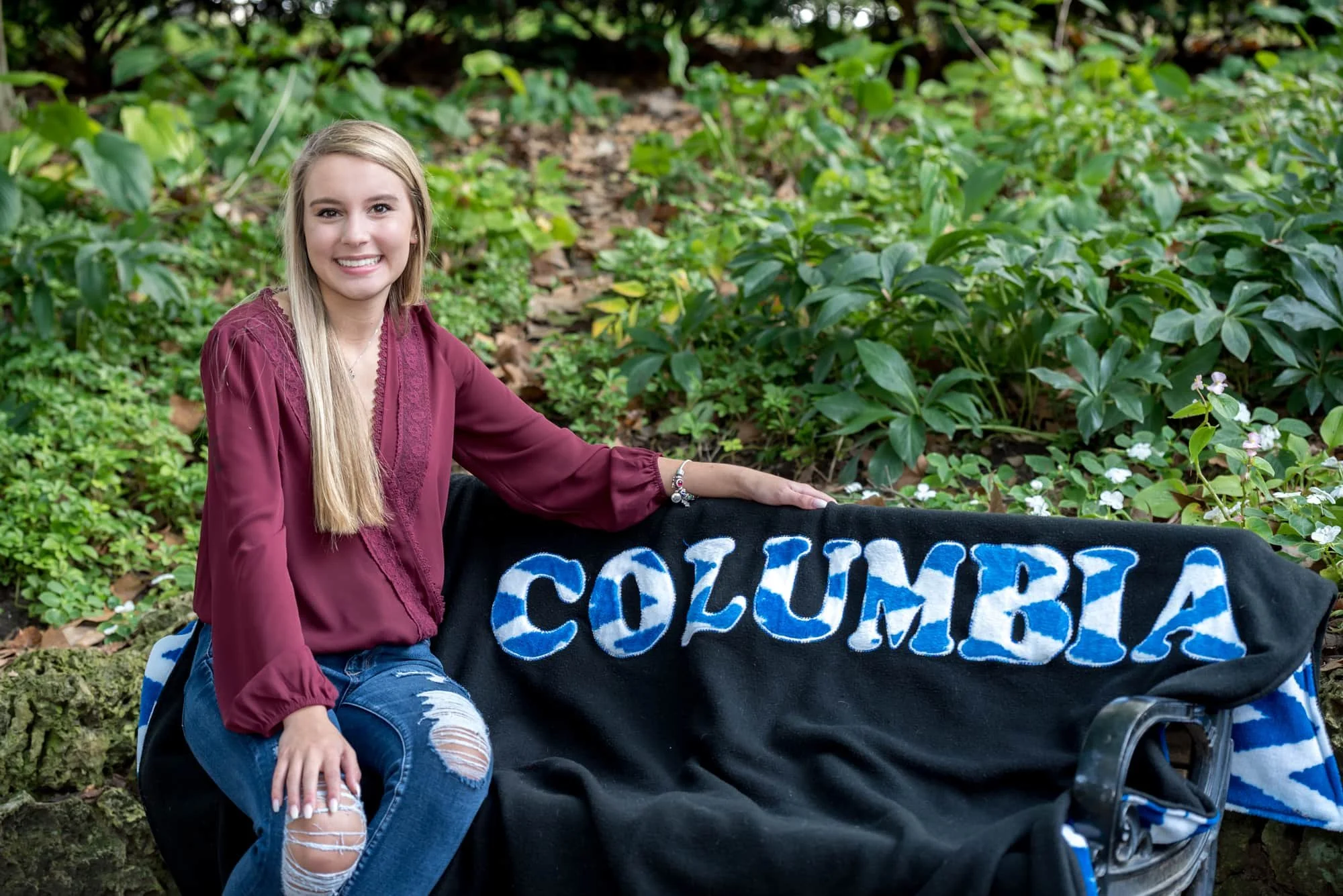 Cozy senior picture of a girl wearing a maroon long-sleeve blouse and ripped jeans, sitting on a park bench at Lafayette Park with a custom embroidered high school spirit blanket laid out beside her.