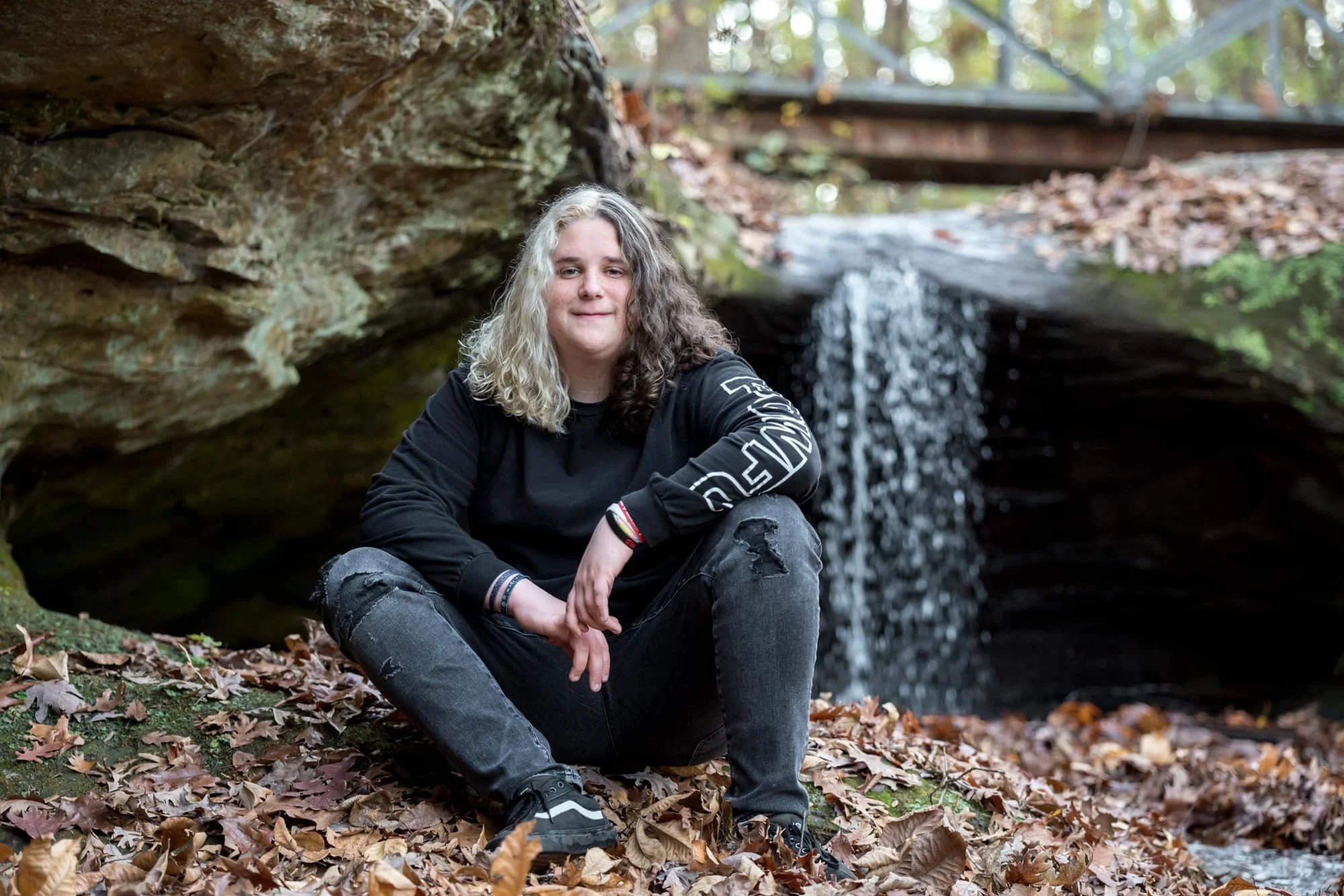 Moody alternative senior photo taken by a Monroe County senior photographer, featuring a boy with half-white and half-brown hair, wearing a black sweatshirt and black ripped jeans while sitting under a rock overhang near a park creek waterfall.