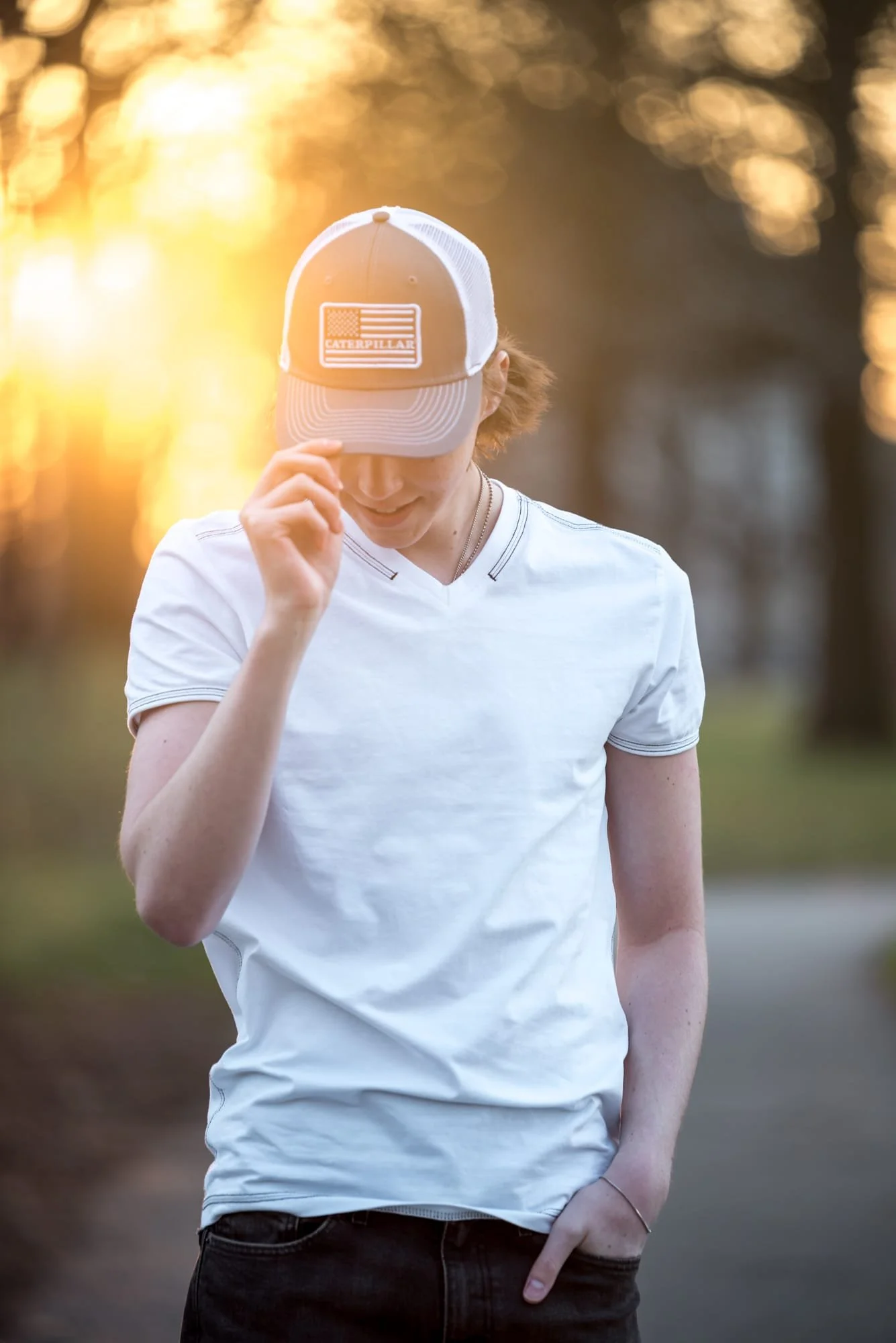 Patriotic sunset senior photo capturing a boy in a white t-shirt and black jeans pulling down the brim of his baseball cap to showcase an American flag patch on the hat at Lafayette Park.