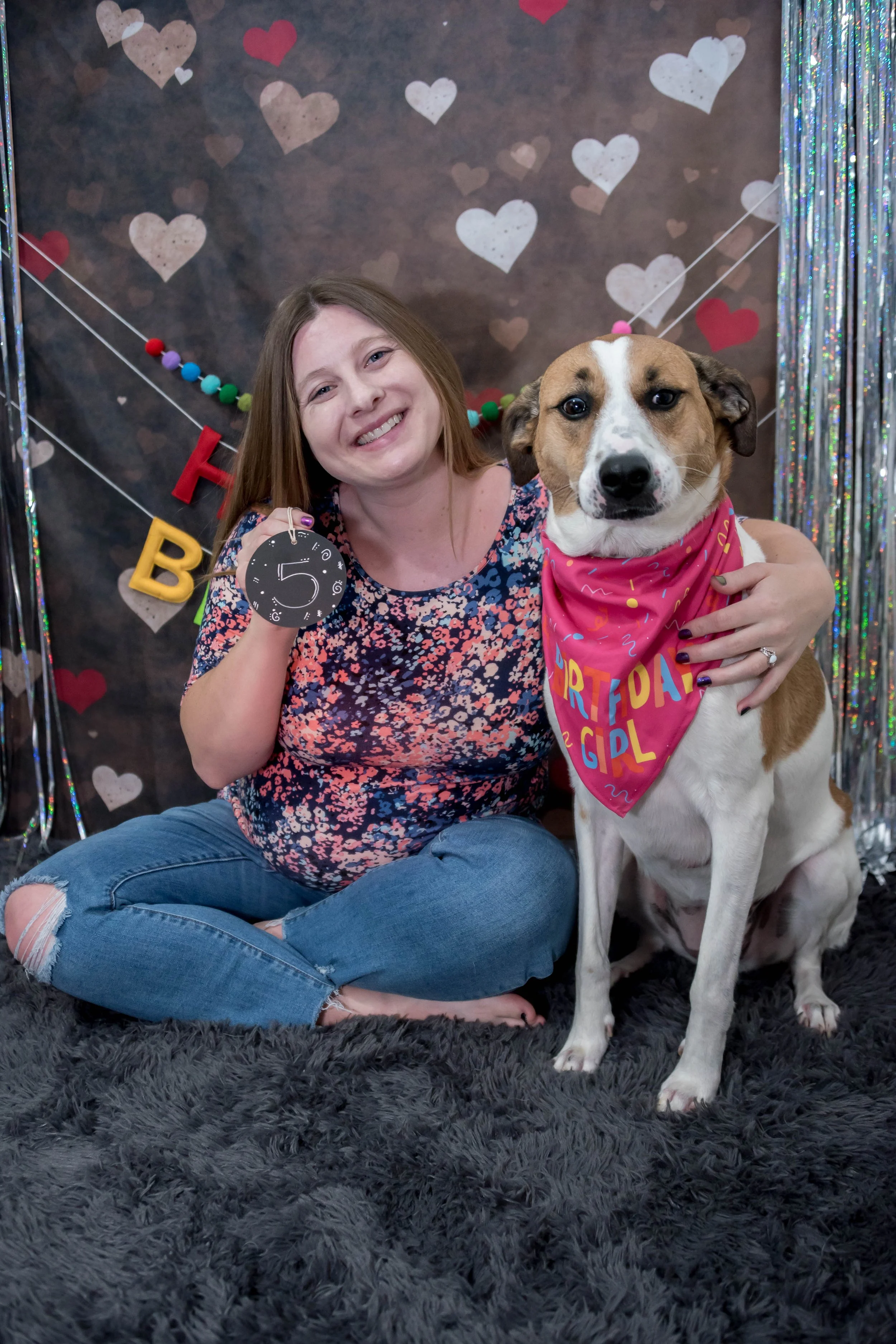 Studio pet portrait featuring a photographer barefoot in a floral top and ripped jeans with her arm around her hound mix dog against a "Happy Birthday" and sparkly streamer and heart background on a grey fuzzy carpet.