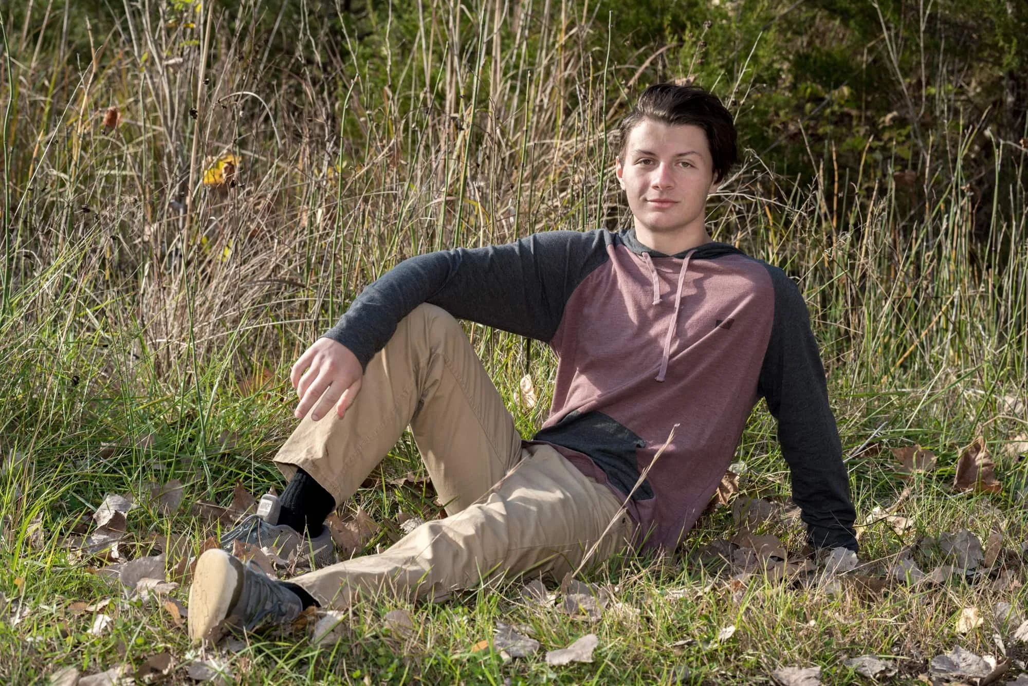 Edgy senior picture of a guy wearing a grey and red hoodie, khaki pants, and tennis shoes, leaning back with his arm on his knee while sitting in the grass at Rock City in Valmeyer IL.
