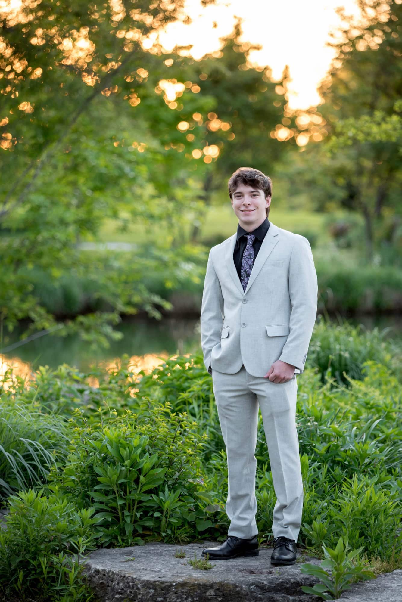 High school senior boy in a light grey suit and paisley tie standing in tall green grass by a creek in Forest Park.