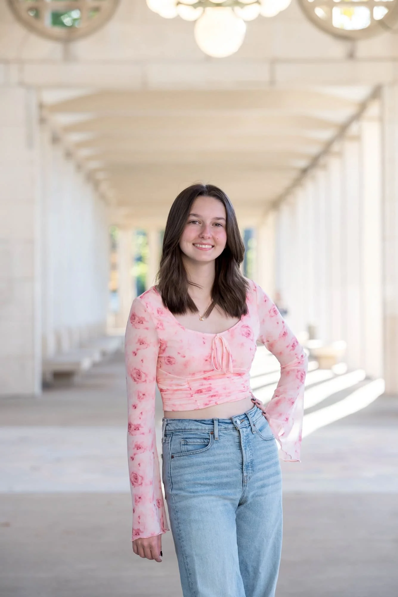 Standing on a concrete walkway at The Muny in St. Louis, a high school senior girl poses with one hand on her hip for an urban style photo wearing a pink floral crop top with blue jeans.