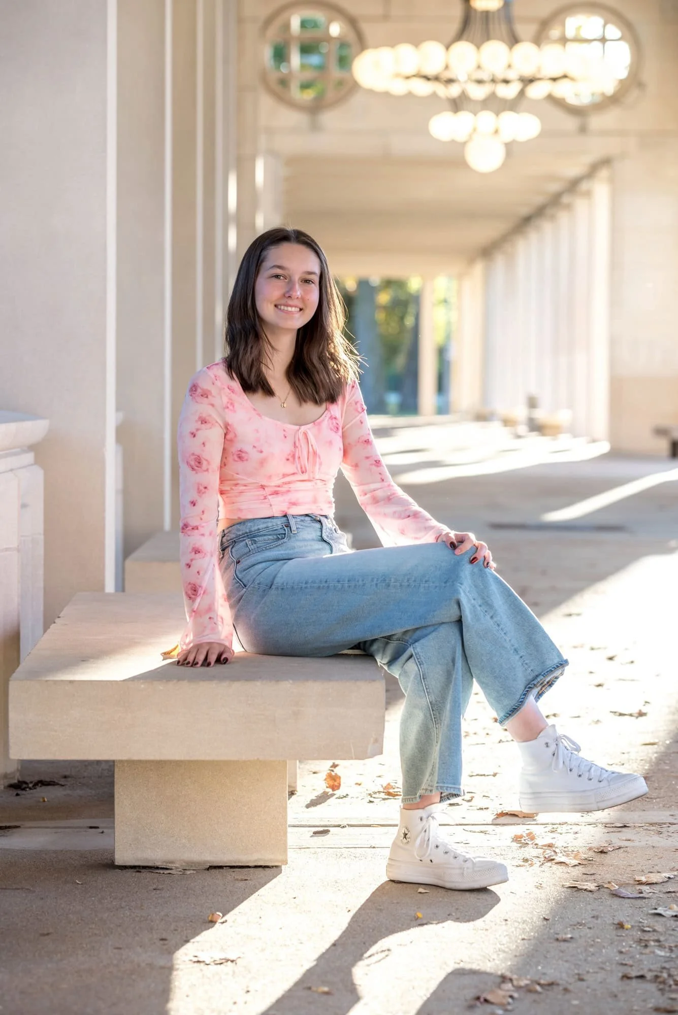 On a park bench at The Muny in St. Louis, a smiling senior girl poses in a pink floral crop top, blue jeans, and white shoes.