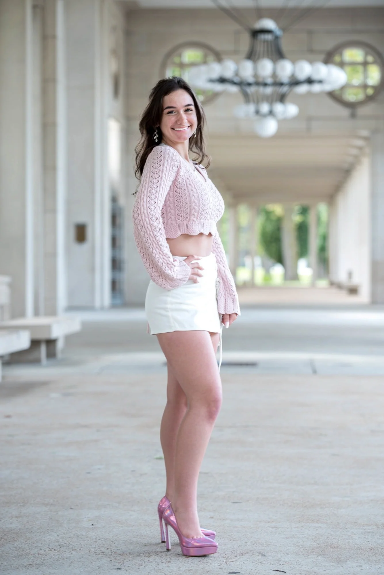 Senior girl wearing a pink crop top, white skirt, and shiny pink heels, standing with her hand on her hip near the iconic chandelier at The Muny in Forest Park.