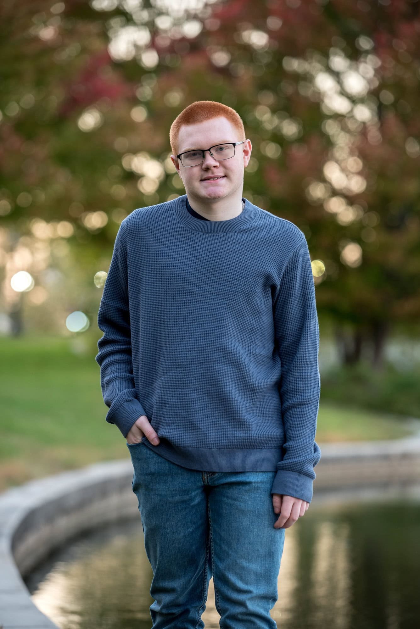 Vibrant autumn senior photo at The Muny  capturing a boy wearing a blue long-sleeve shirt and blue jeans in front of a scenic lake.