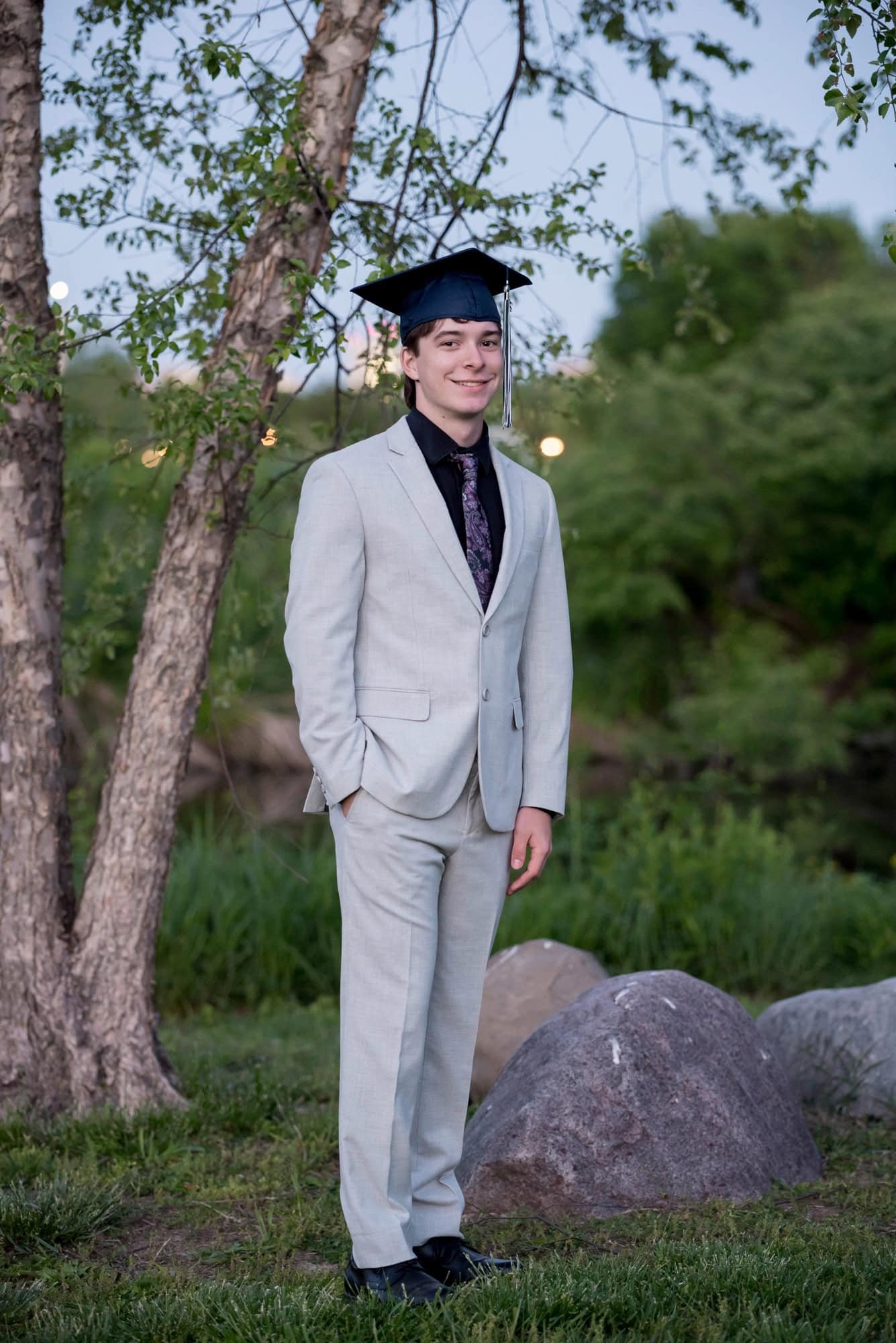 High school senior boy wearing a graduation cap with a formal light grey suit standing next to a tree in Forest Park.