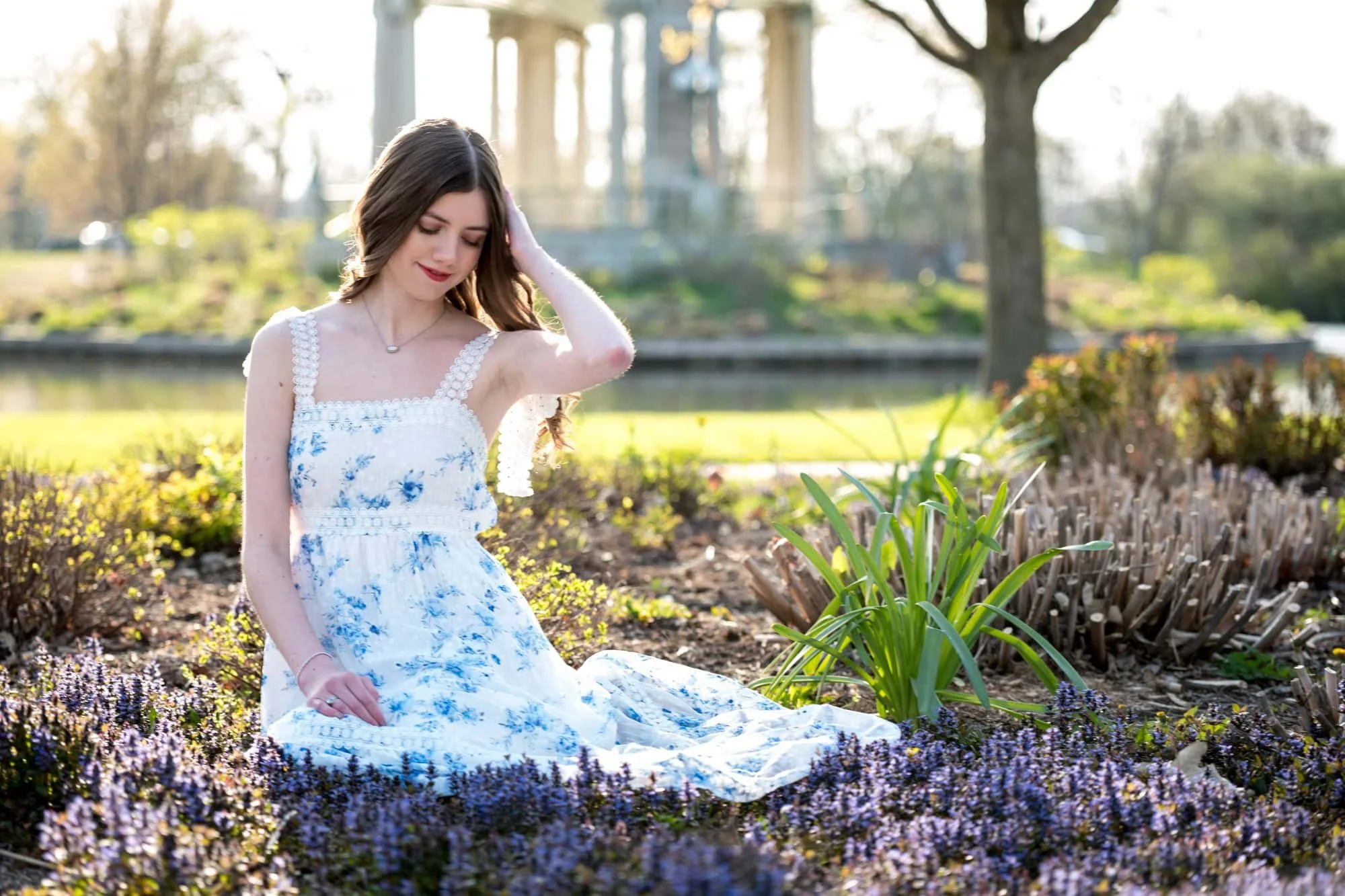 High school senior photo of a girl sitting in a lush flower patch in front of the historic Muny pagoda in Forest Park, wearing a white dress with blue flowers for a dreamy, fairytale look.