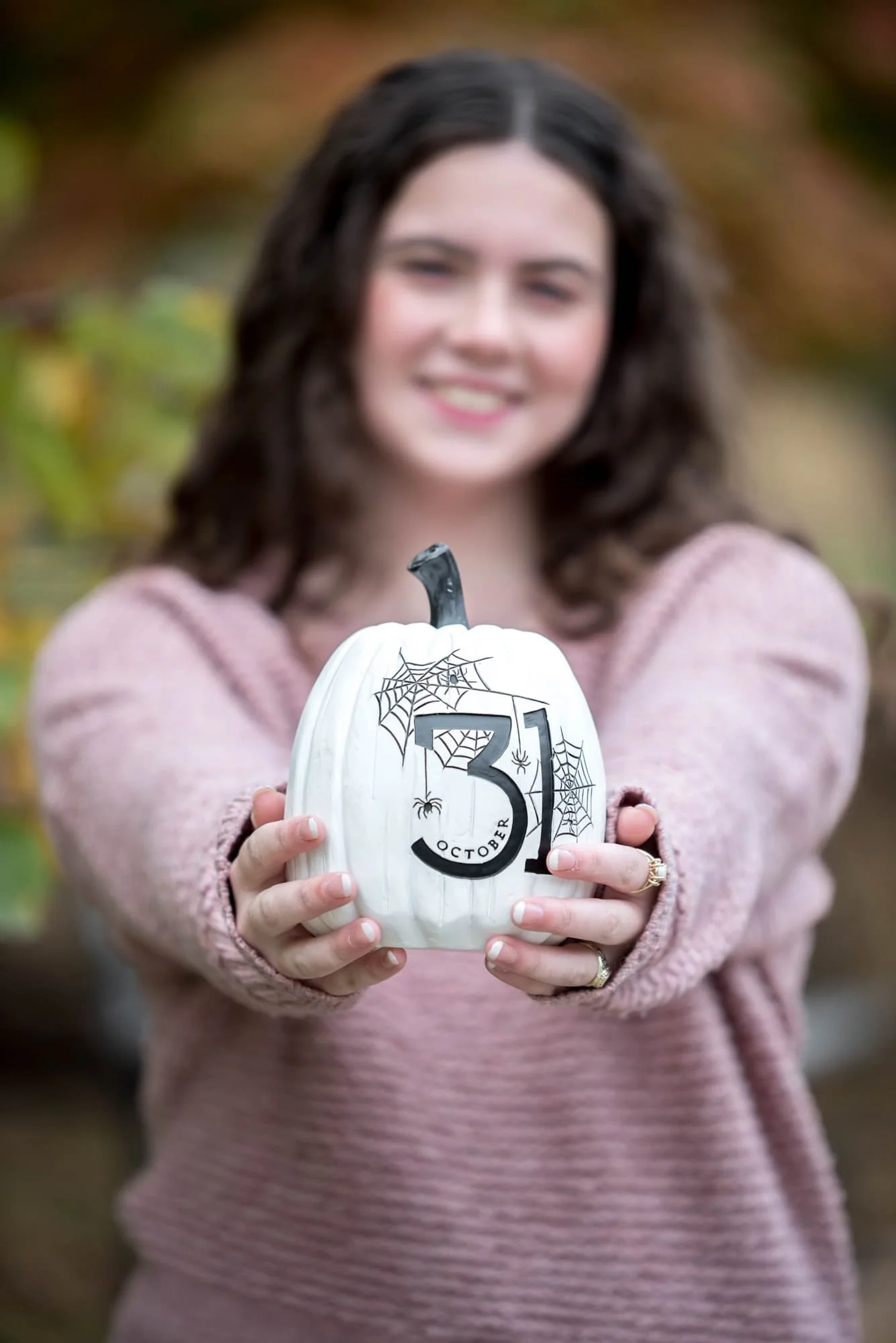 A fall session photo captures a senior girl in a pink sweater and blue jeans holding a pumpkin in St. Louis, MO.