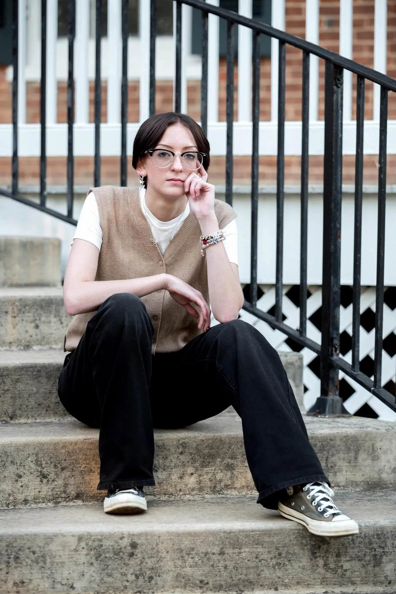 Moody park senior photo of a girl with a serious expression sitting on the curved brick staircase at the back of the Victorian Jarville House at Queeny Park, wearing a tan vest and white t-shirt.