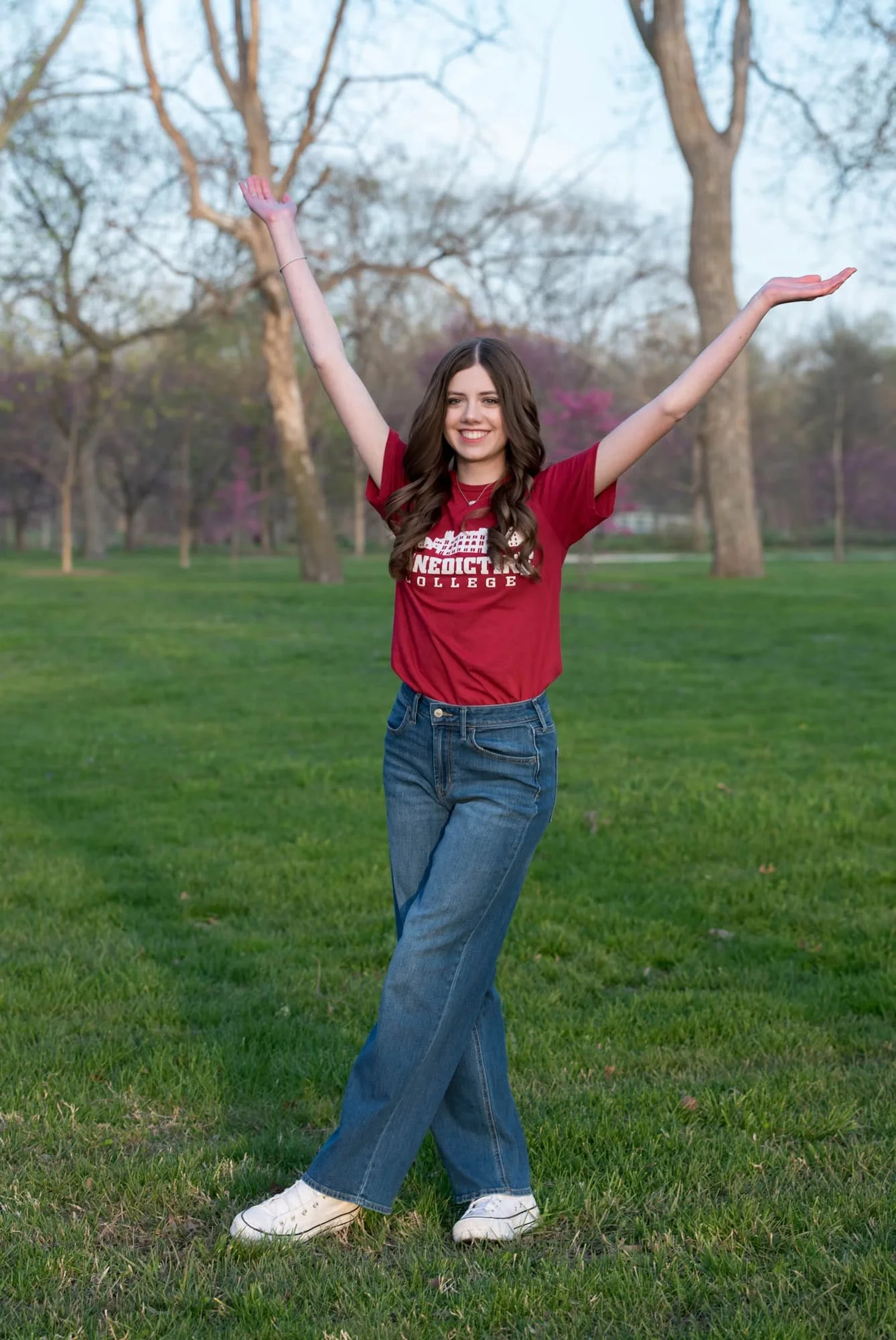 Senior photo of a girl wearing a red college shirt, blue jeans, and white Converse sneakers, standing in an open spring grassy field with her hands in the air and her feet crossed at Forest Park in St. Louis.