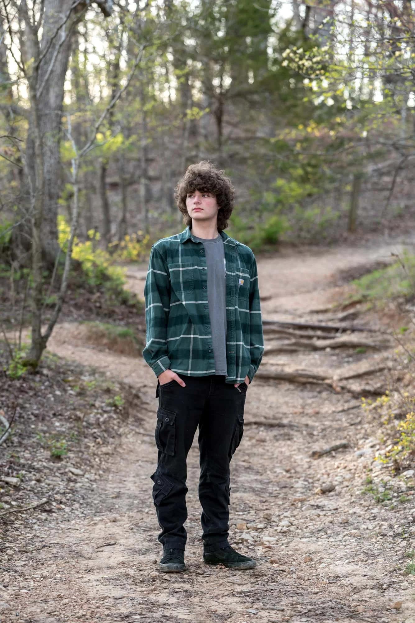 Senior photo of a guy standing on a wooded pathway with sunlight peeking through the early spring trees, wearing a green plaid long-sleeve and black pants at Castlewood State Park in Ballwin.