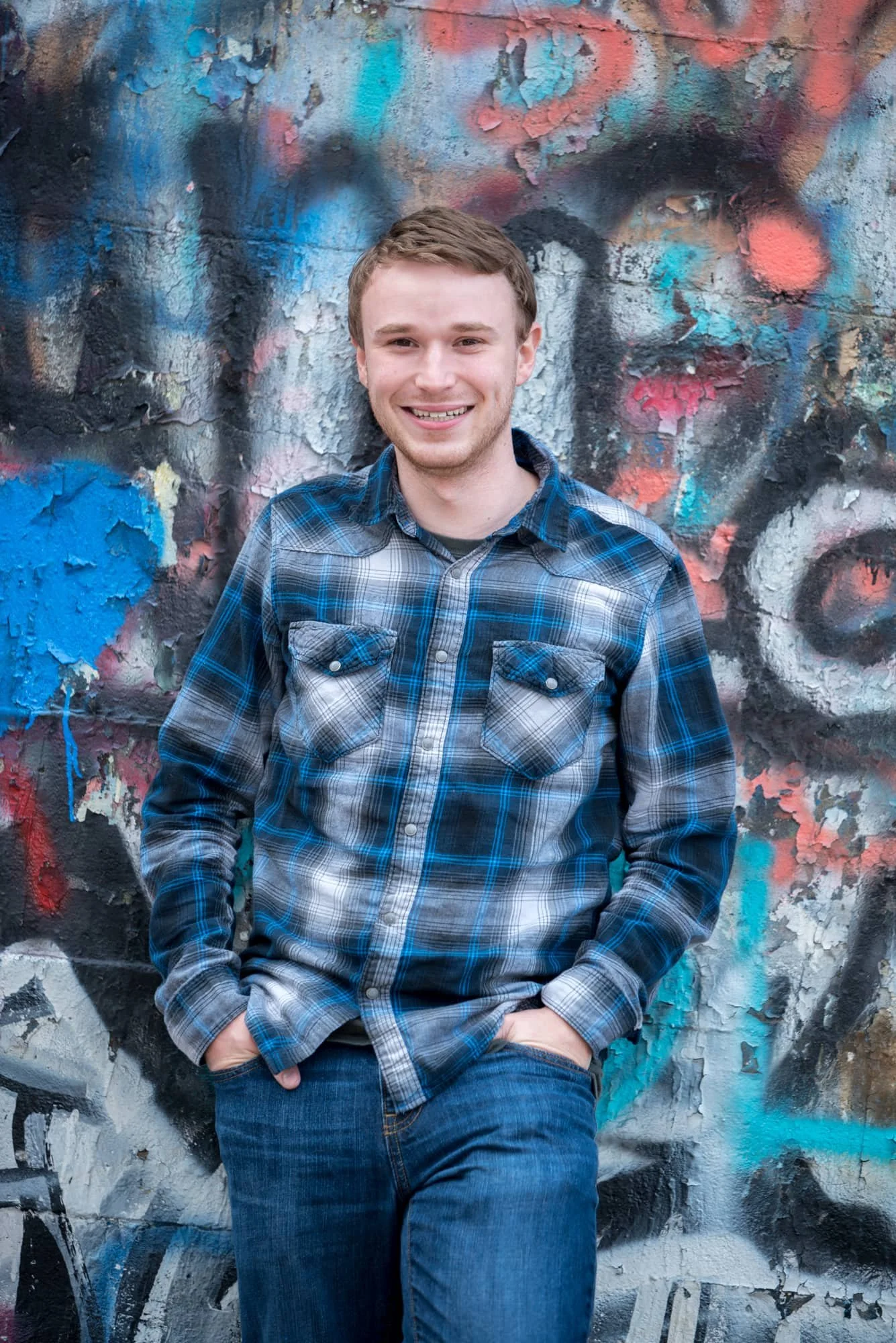 Edgy urban senior portrait of a guy standing with his hands in his pockets in front of a colorful graffiti wall in Columbia IL, wearing a classic blue plaid long-sleeve shirt and blue jeans.